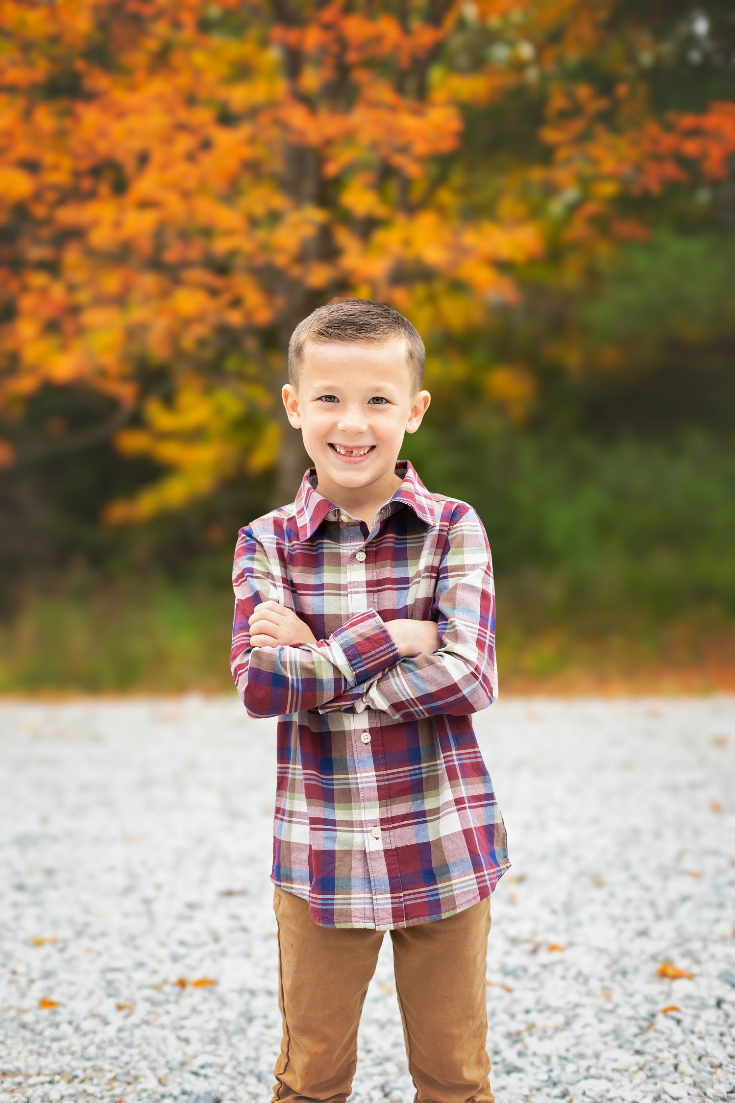 A smiling boy in a plaid shirt stands with arms crossed on a gravel path with fall foliage behind.