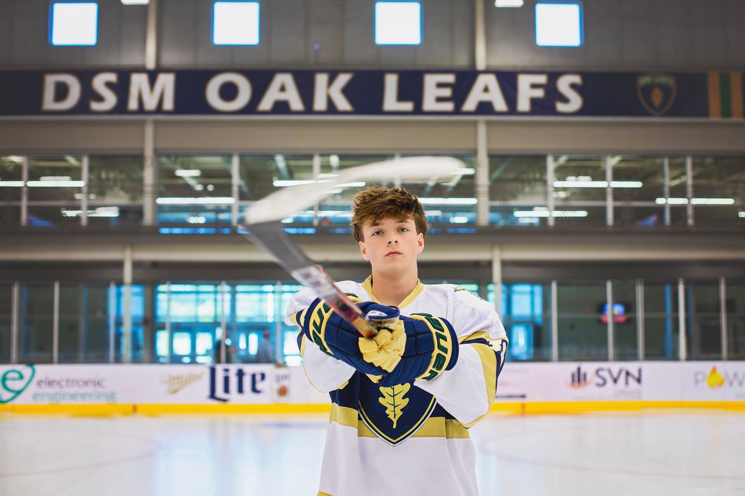 Teen hockey player in DSM Oak Leafs uniform holding stick on ice rink.