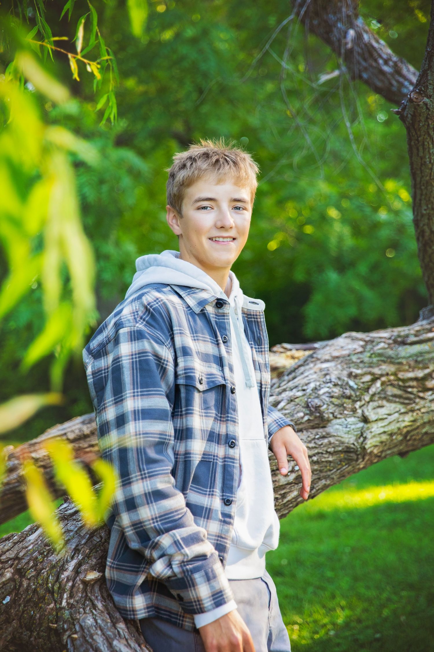 Smiling teenage boy in plaid shirt leaning against tree trunk in sunny green park setting.