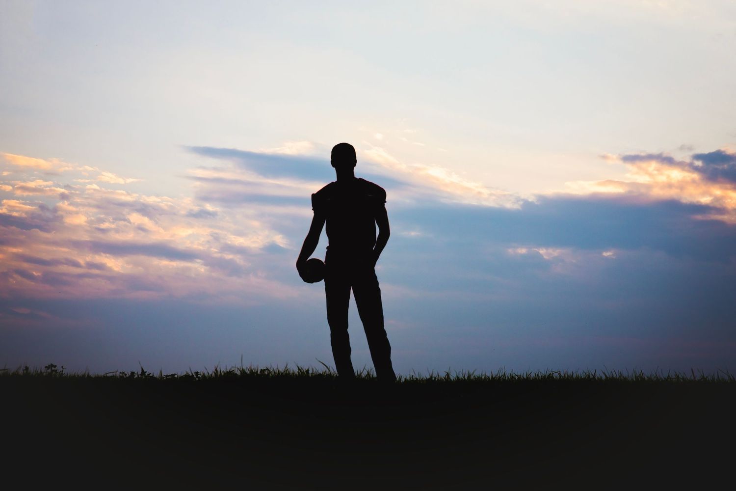 Silhouette of a man holding a football standing on a grass hill against a dramatic sunset sky.