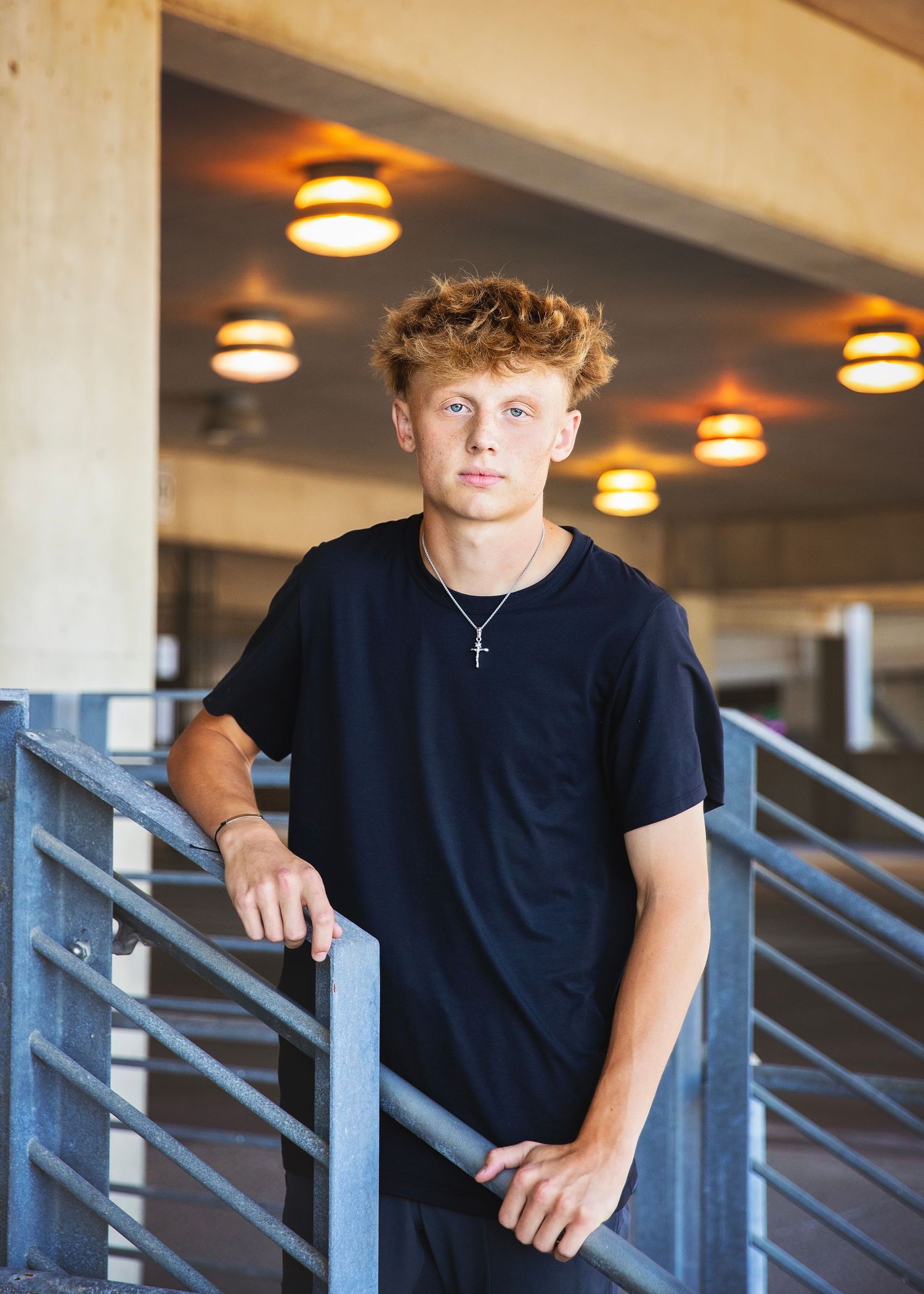 Teen boy with curly red hair leans on metal railing wearing black t-shirt with cross necklace.