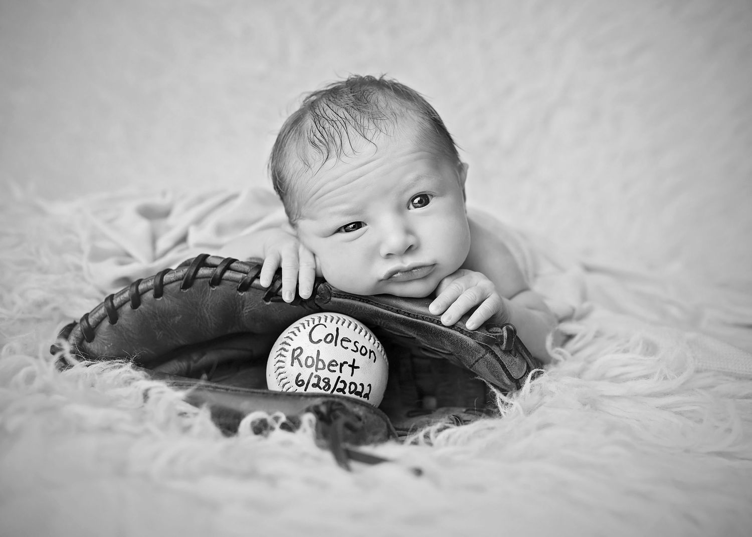 Black and white photo of newborn baby in baseball glove with personalized ball showing name and birthdate.