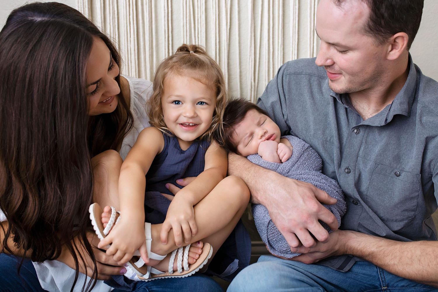 Family of four with toddler girl and parents admiring newborn baby wrapped in blue.