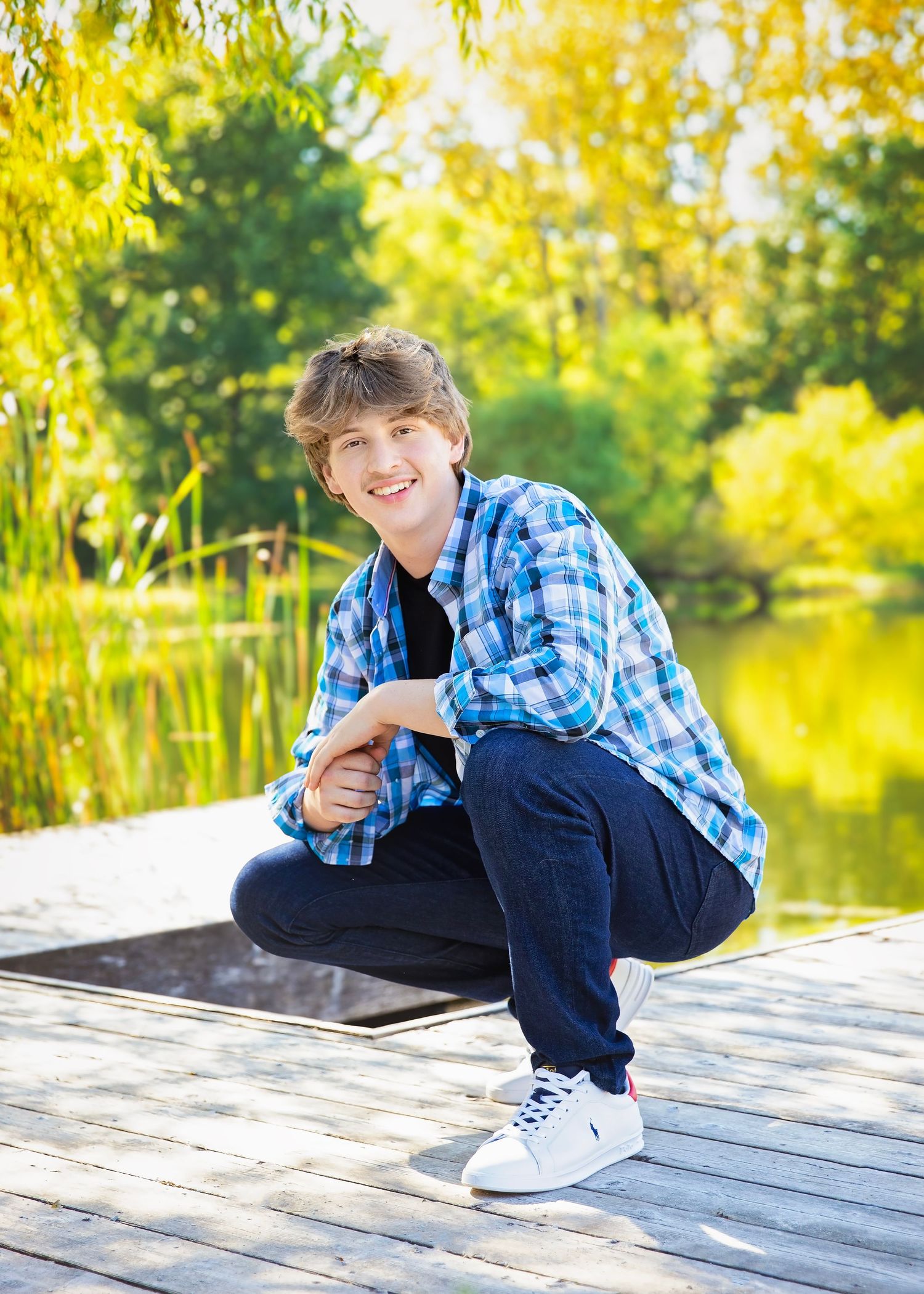 Smiling teen boy in blue plaid shirt crouching on a wooden dock by a sunlit pond for senior photos.