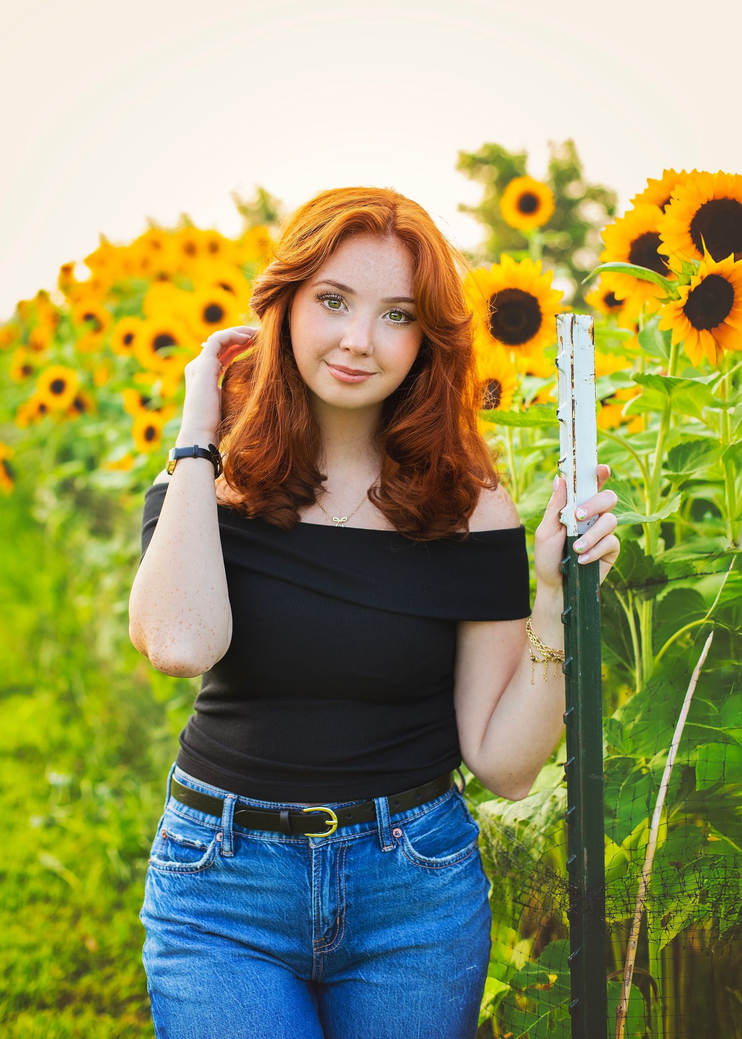 Red-haired girl in black top and jeans posing beside a fence in a vibrant sunflower field.