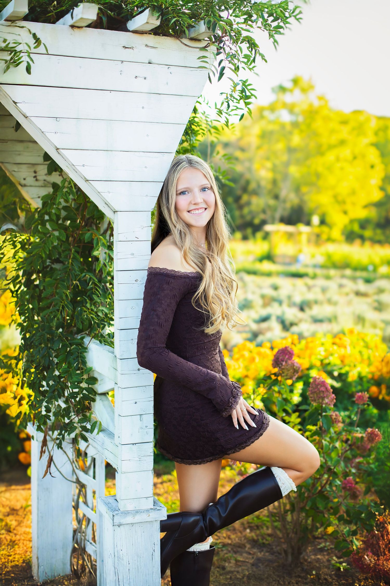 Smiling blonde teen in purple dress and tall boots leans against white garden arbor in flower garden.