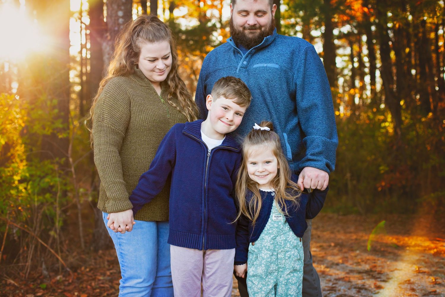 Family of four standing on autumn forest path with golden sunlight filtering through colorful fall trees.