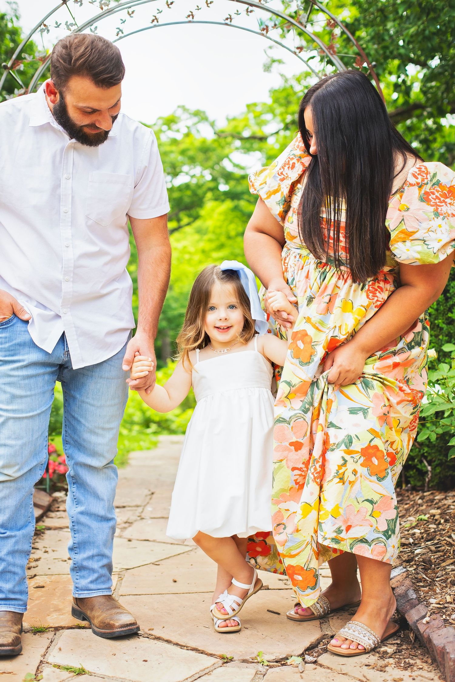 A happy toddler girl in white dress holds hands with parents in a lush garden setting.