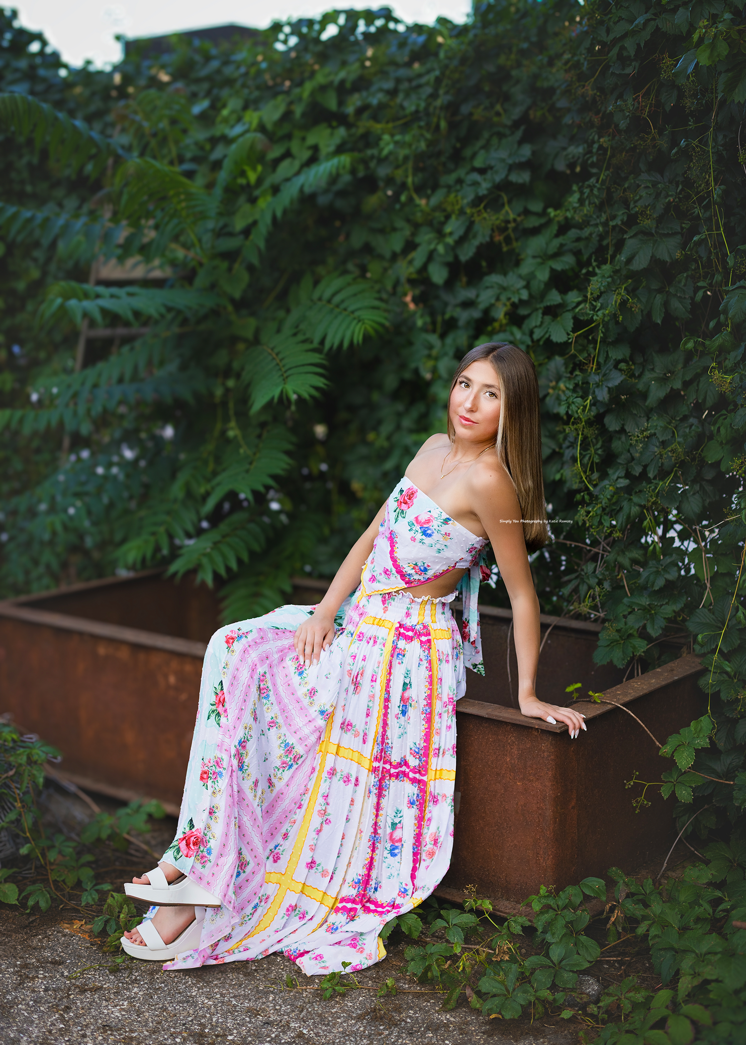 Young woman in floral strapless maxi dress sitting on rustic ledge surrounded by lush green foliage outdoors.