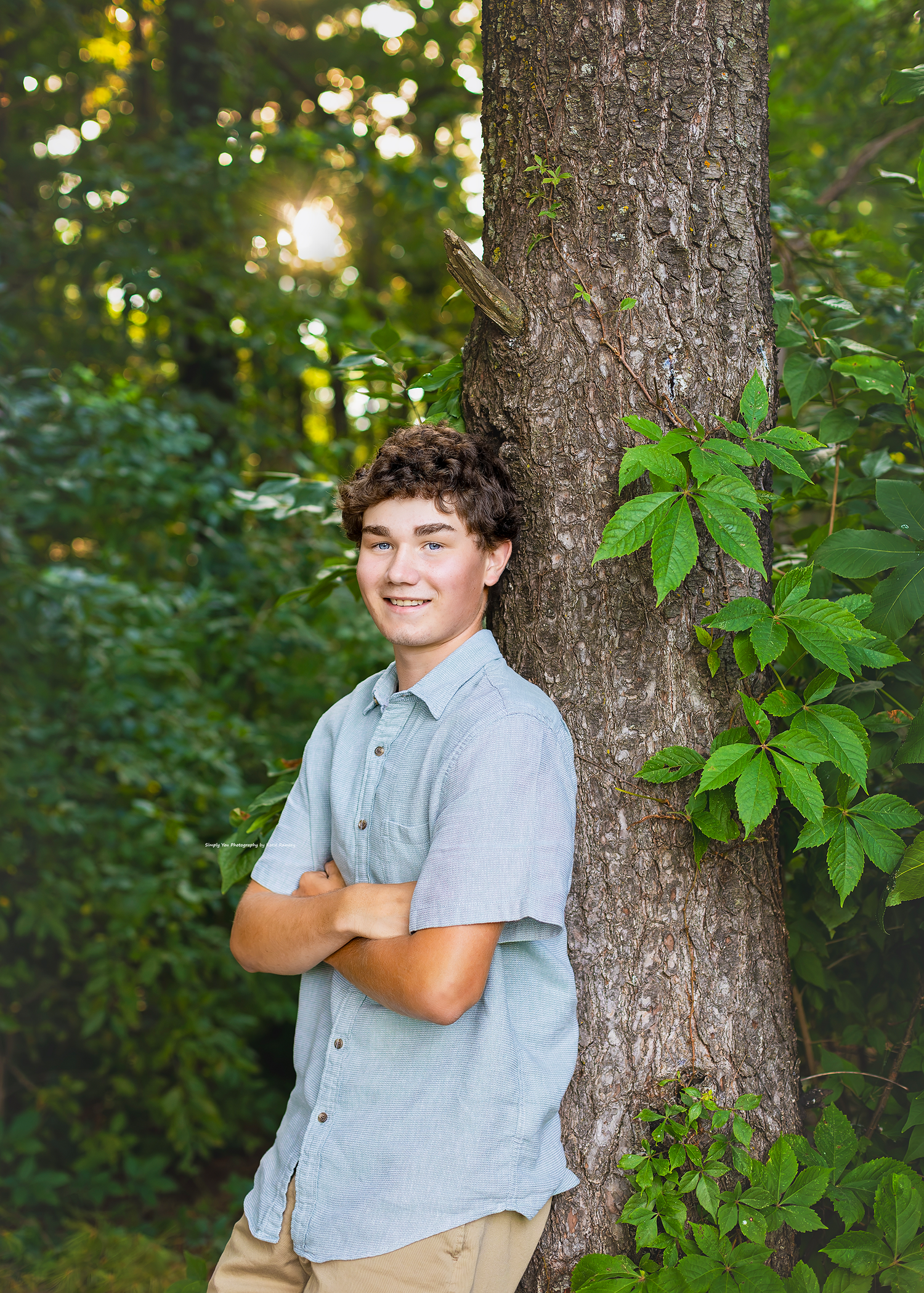 A smiling teenage boy in a blue shirt leans against a tree trunk surrounded by lush green foliage outdoors.
