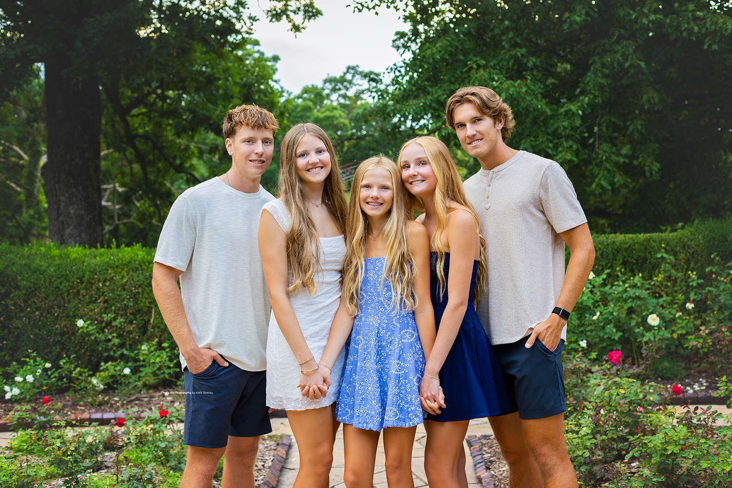 Five smiling teenagers pose together outdoors in a lush garden setting, wearing casual summer outfits.