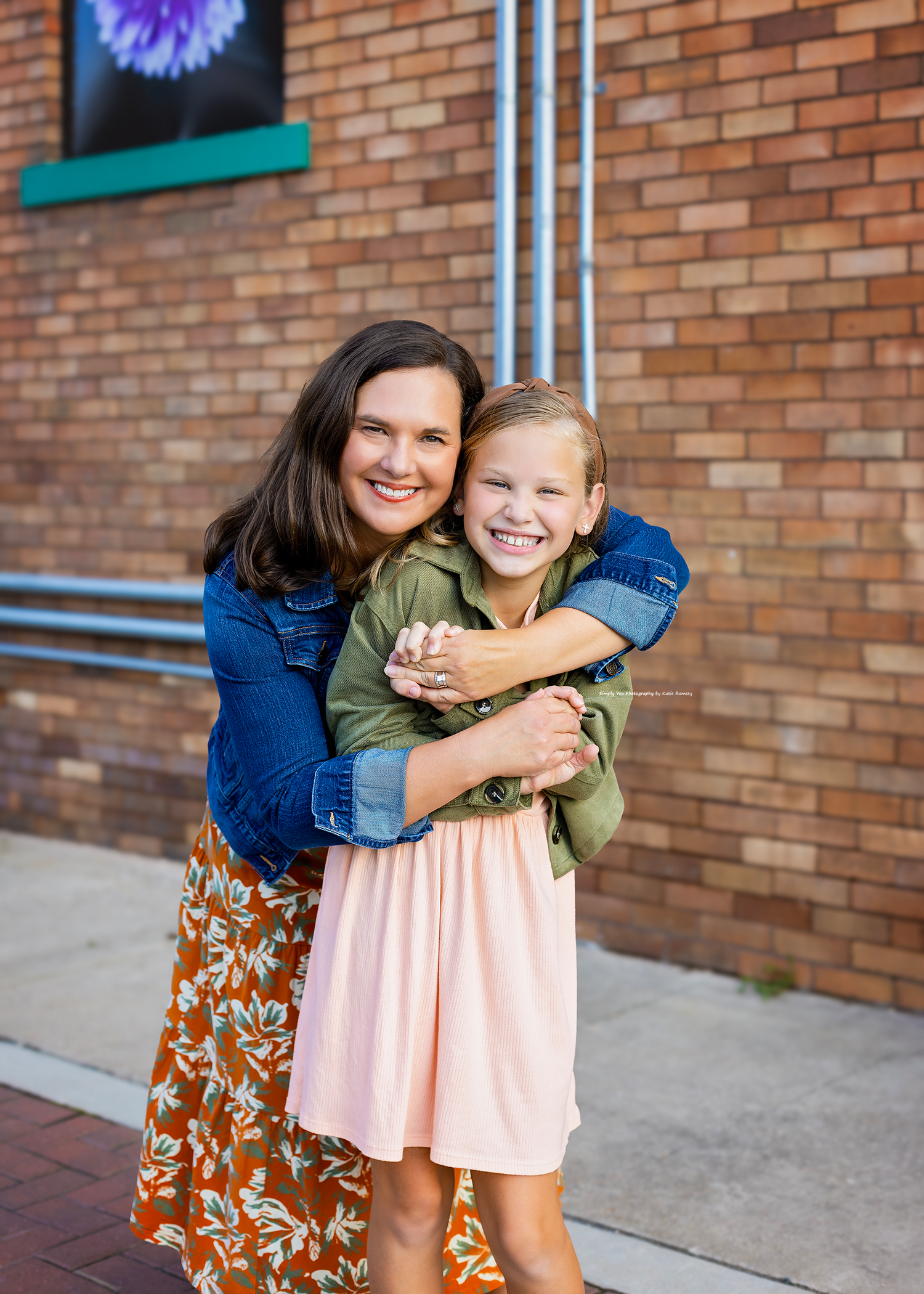 A smiling woman in a floral skirt hugs a young girl in a pink dress and denim jacket in front of a brick wall.