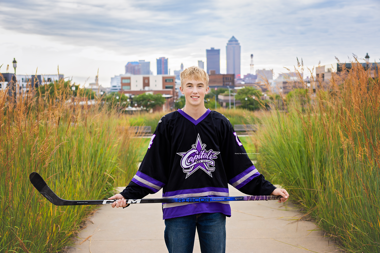 Teen boy in hockey jersey holds stick on path with city skyline and tall grass in background.