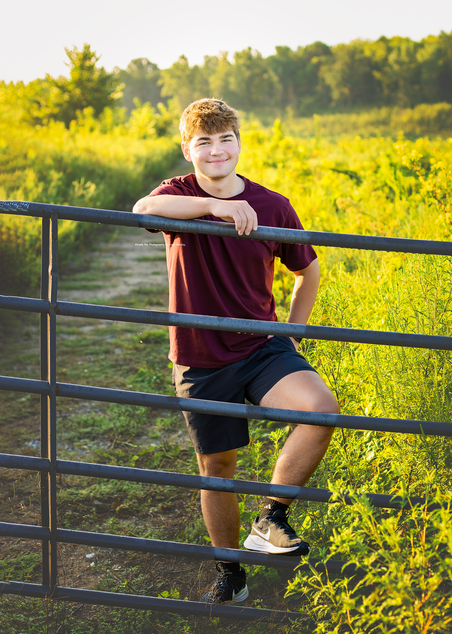 Teen boy in maroon shirt leans on metal farm gate, smiling outdoors in sunny green field.