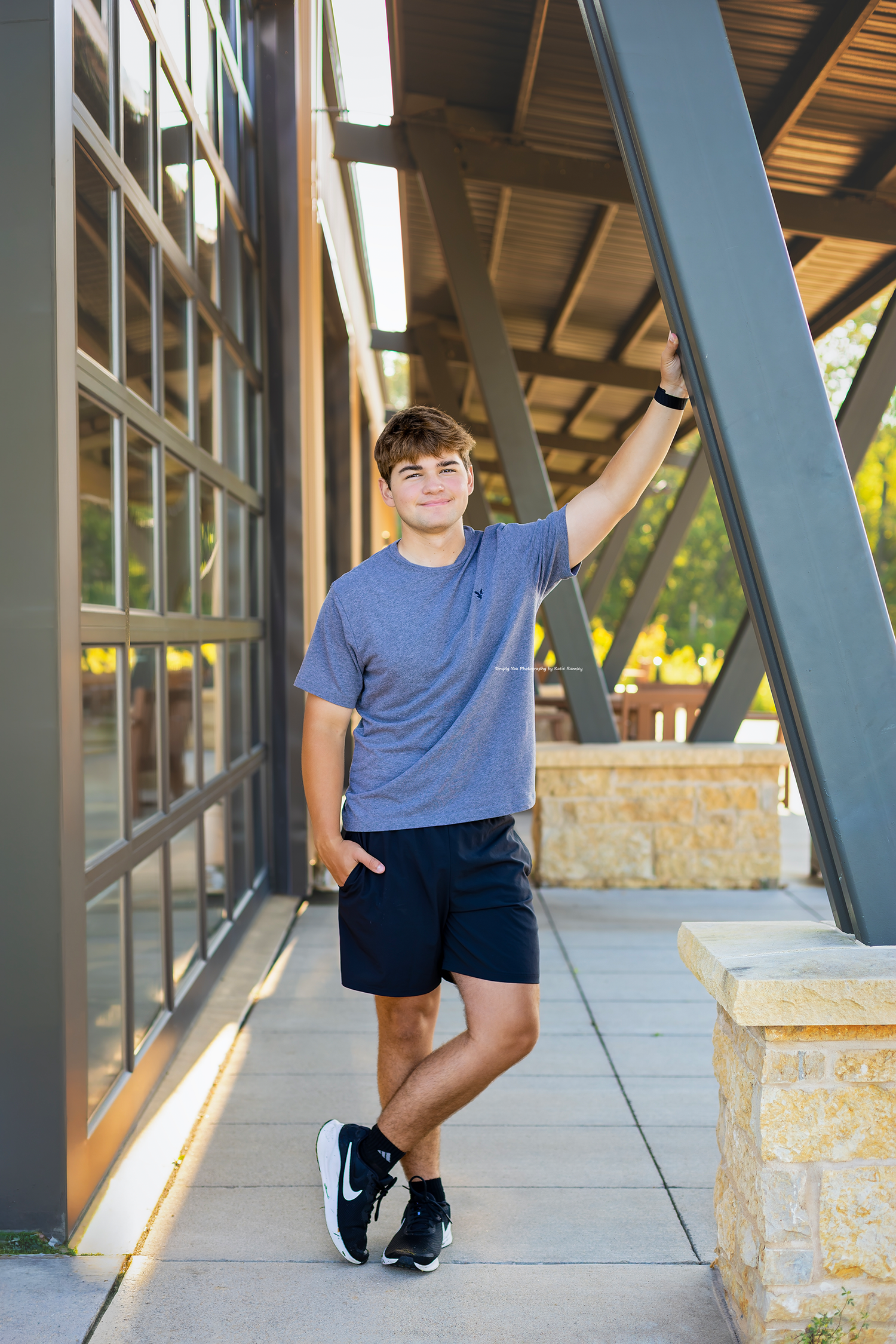A young man in a blue t-shirt and navy shorts leans against a modern building structure, smiling outdoors.