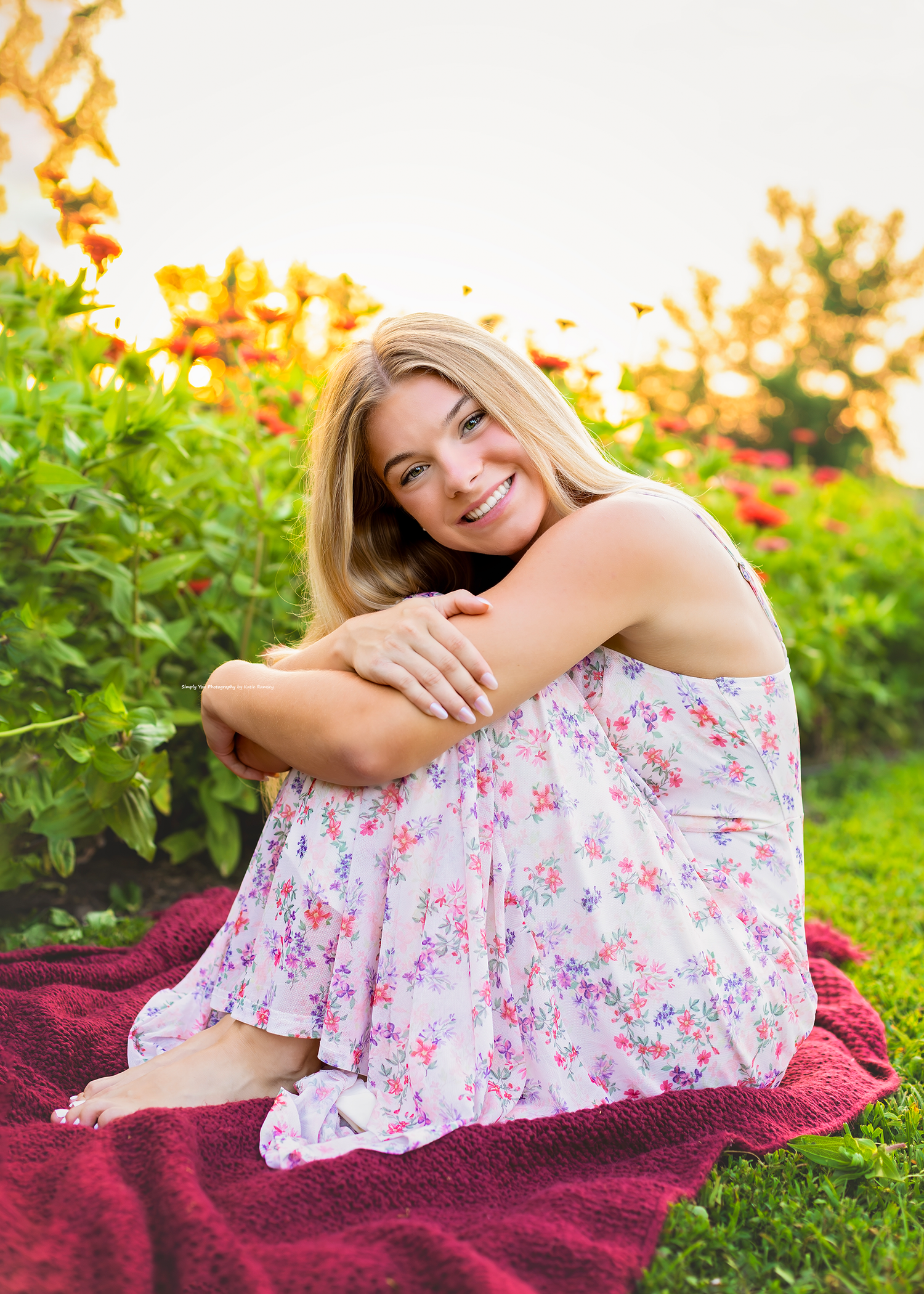 Young blonde woman in floral dress sitting on red blanket surrounded by colorful garden flowers, smiling outdoors.