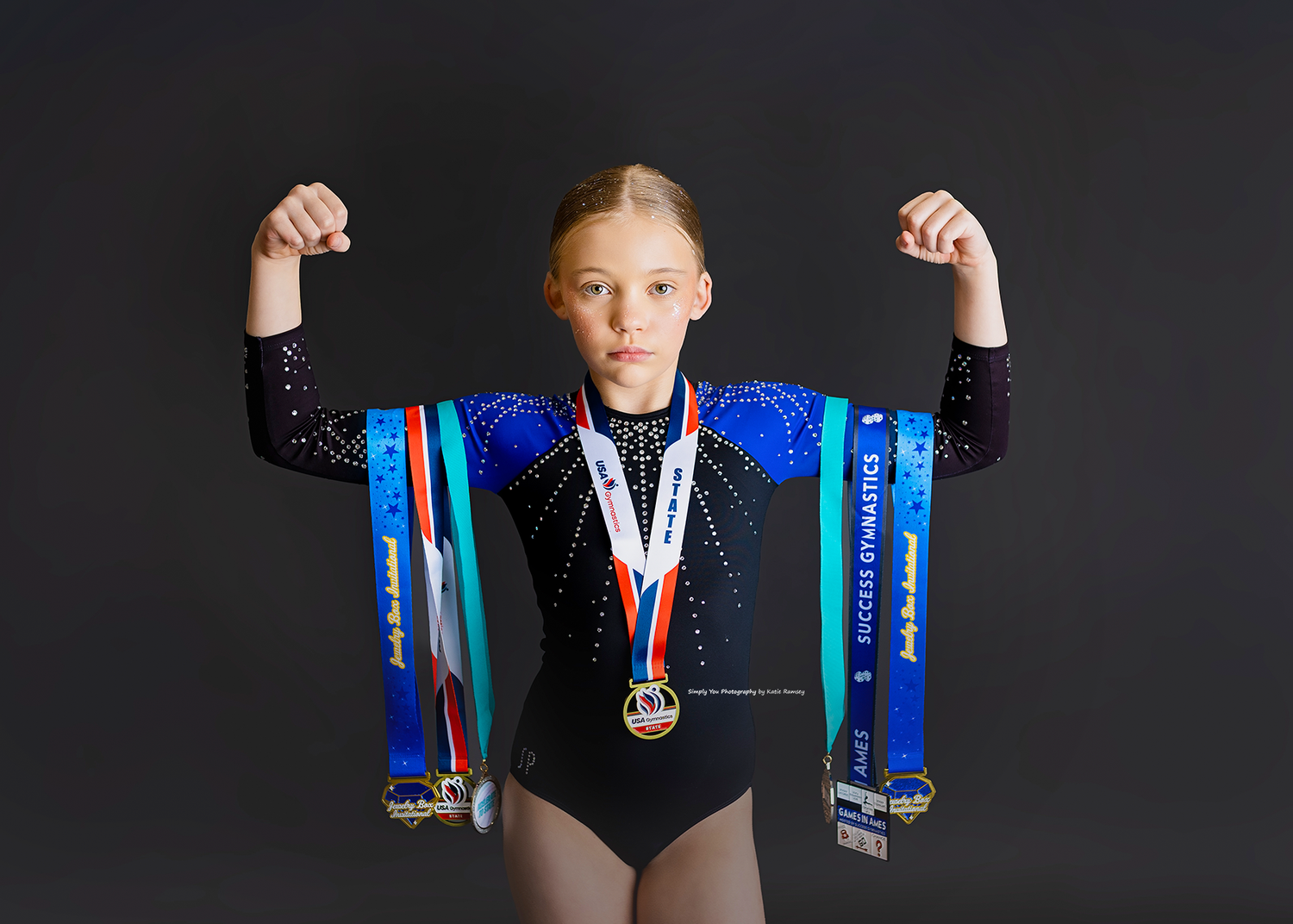 Young gymnast flexing with multiple medals and colorful ribbons in a black leotard against dark background.
