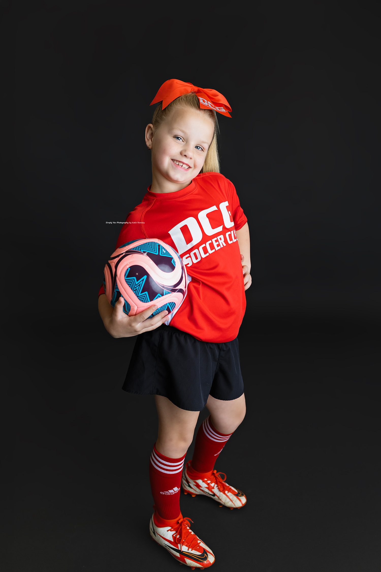 Young girl in red DCC Soccer Club jersey, black shorts, red Adidas cleats, and orange bow holds a soccer ball.