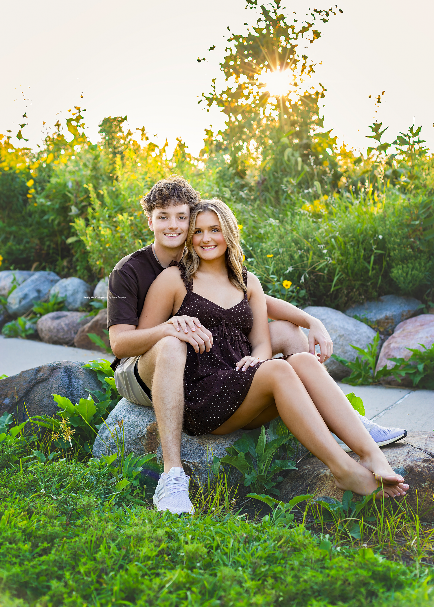 A young couple sits together on rocks in a lush green garden, smiling warmly in golden sunset light.
