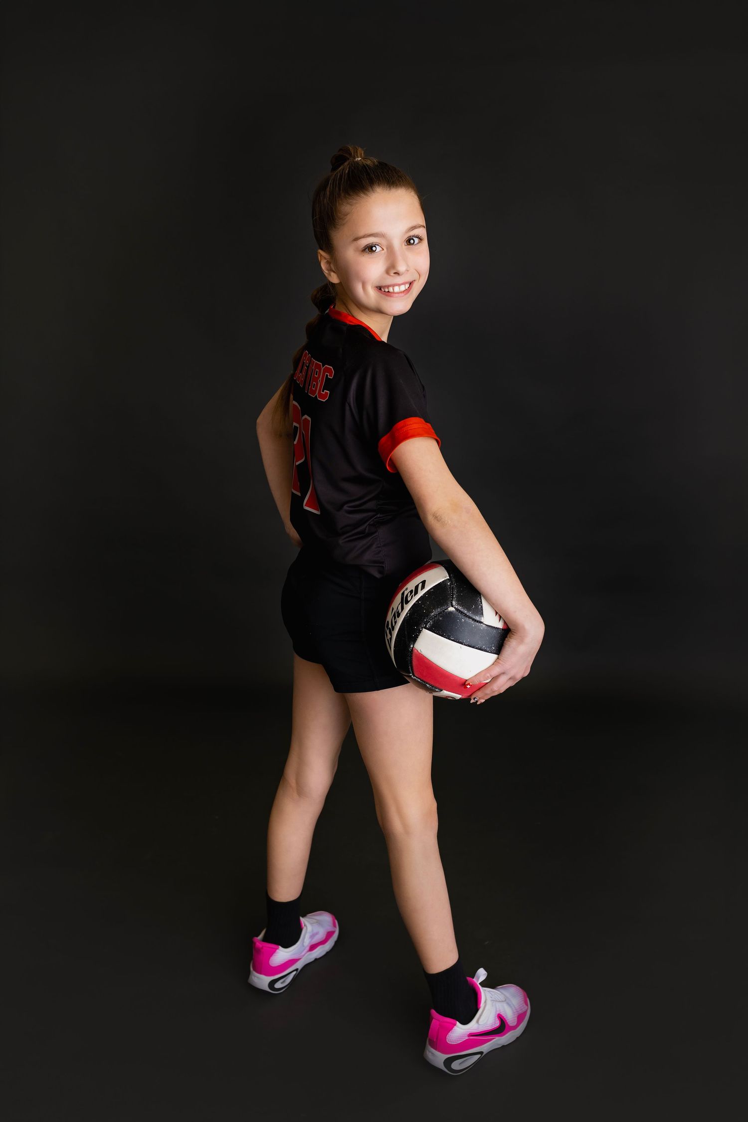 Young girl in black volleyball uniform holding ball standing sideways showing back of jersey in studio.