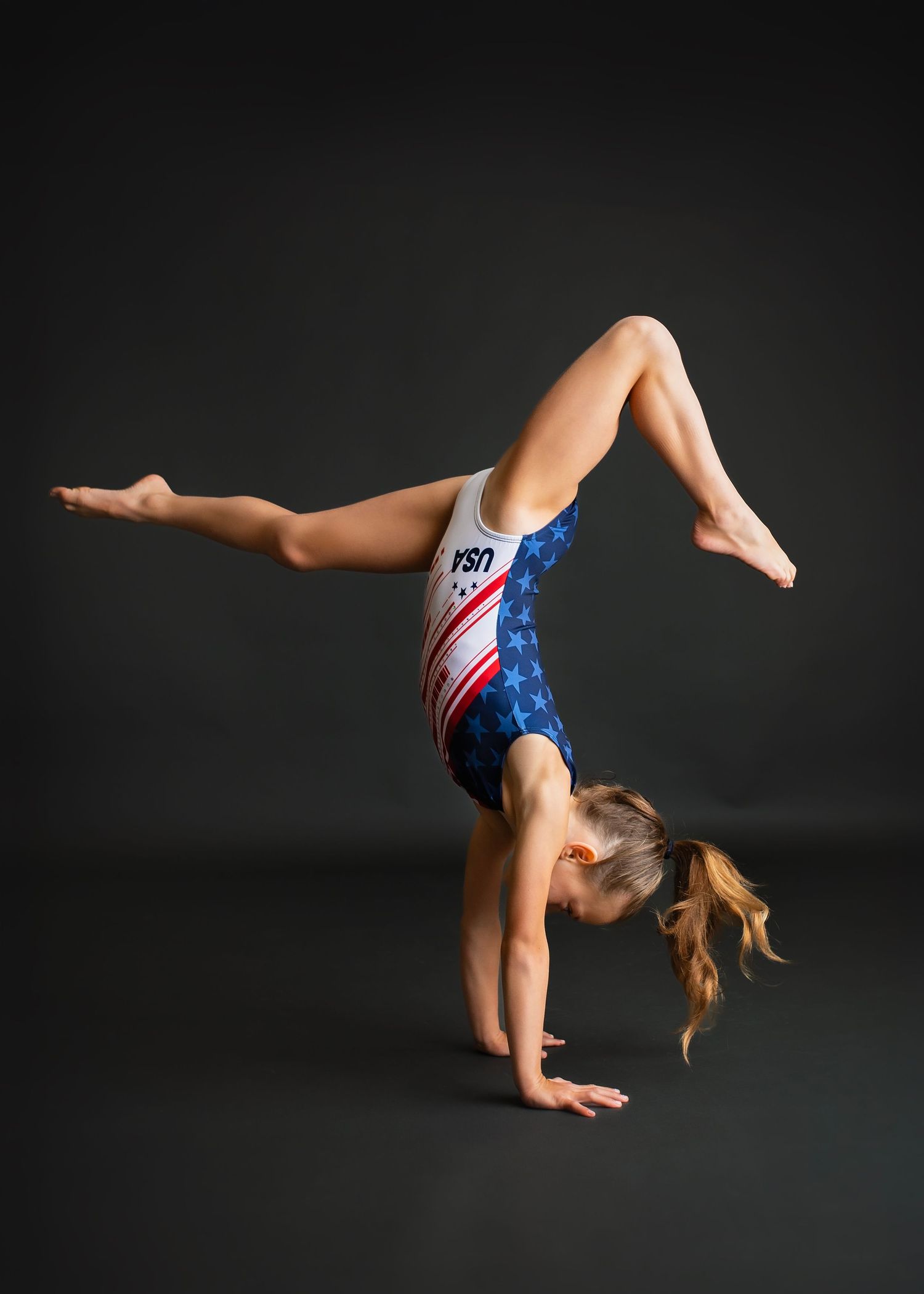 Young gymnast performing a handstand in a USA patriotic leotard against a dark background.