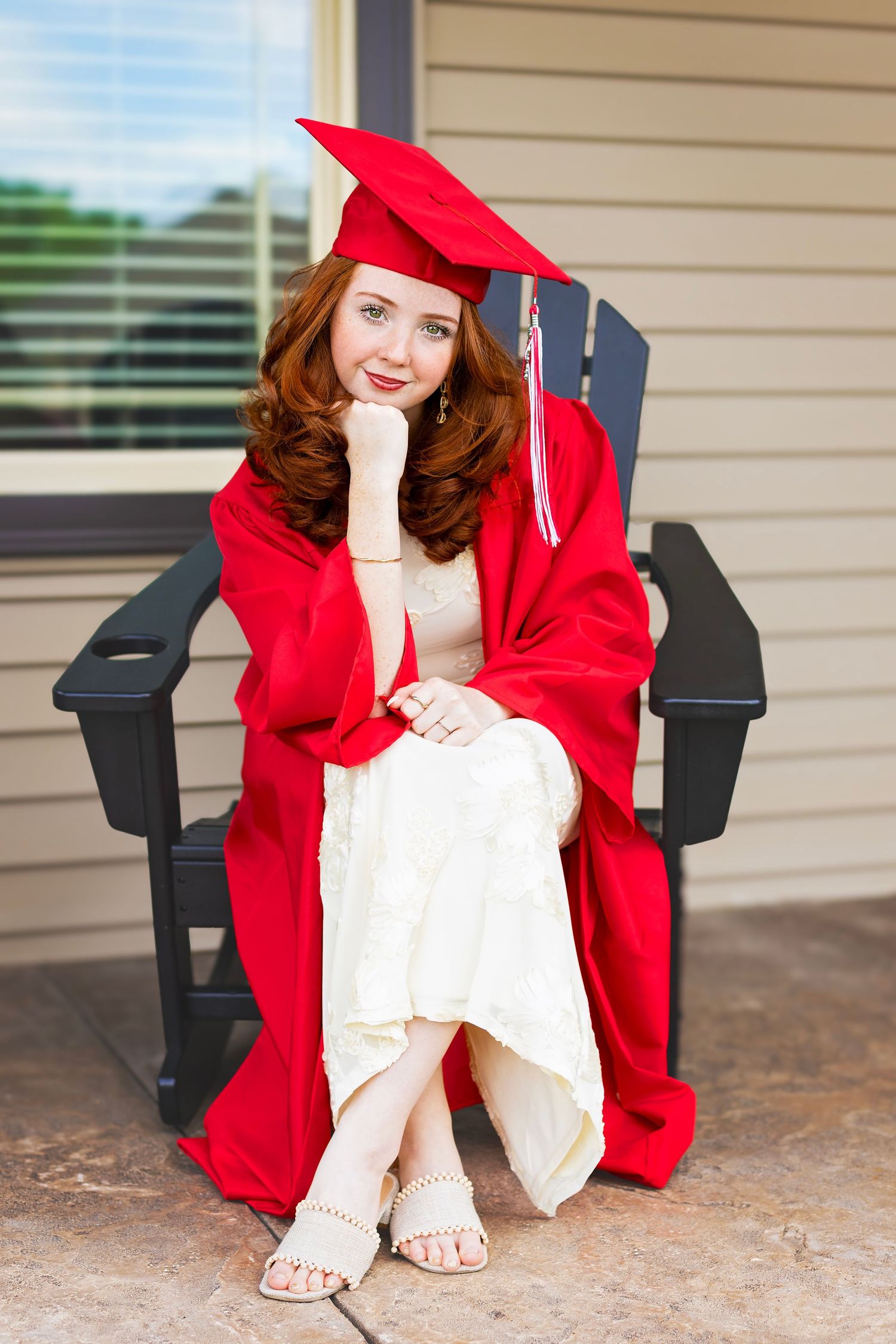 Red-haired graduate in red cap and gown sitting in black chair smiling for senior portrait photo.