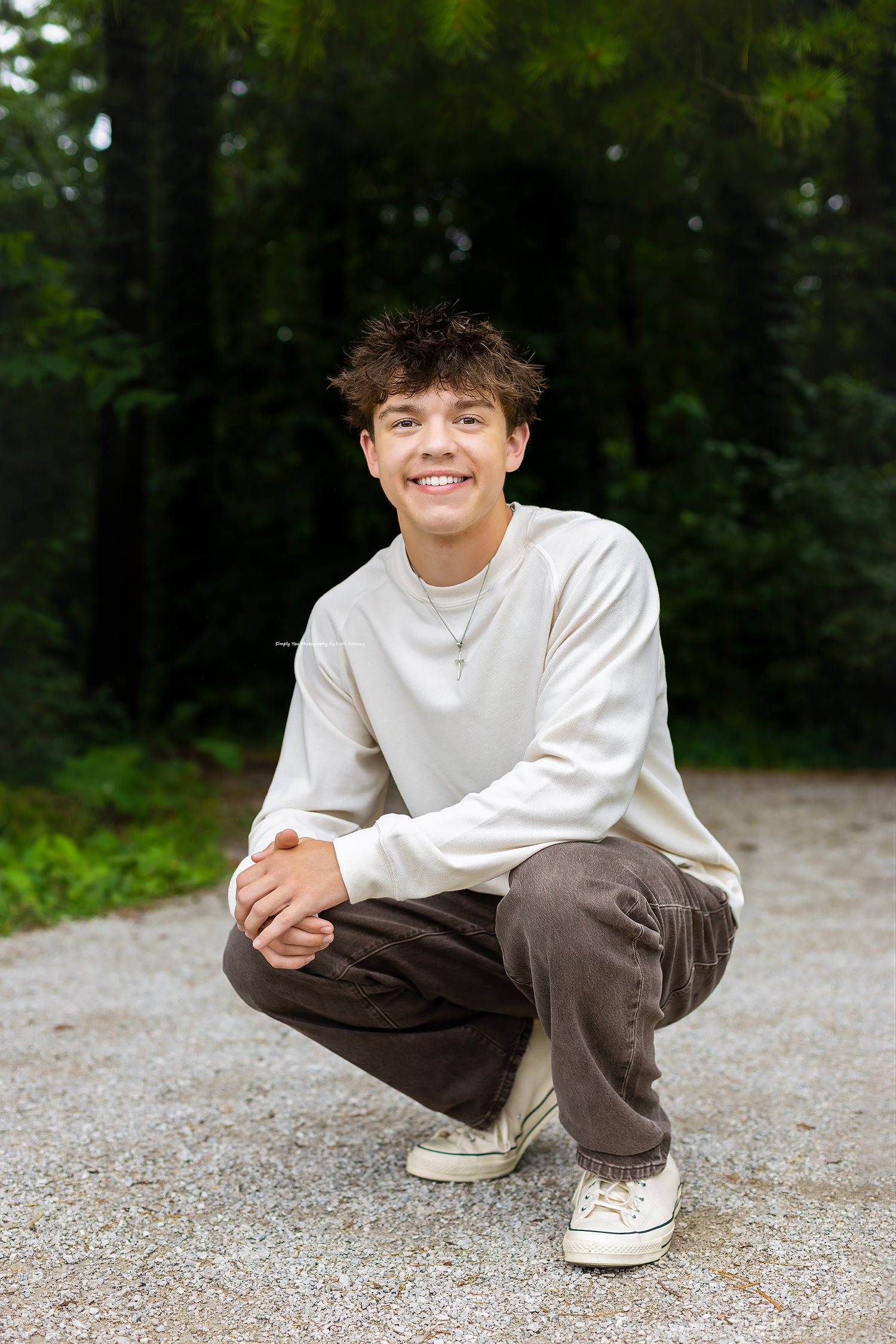 Smiling young man crouching on a gravel path wearing a white sweatshirt and brown pants with white sneakers.