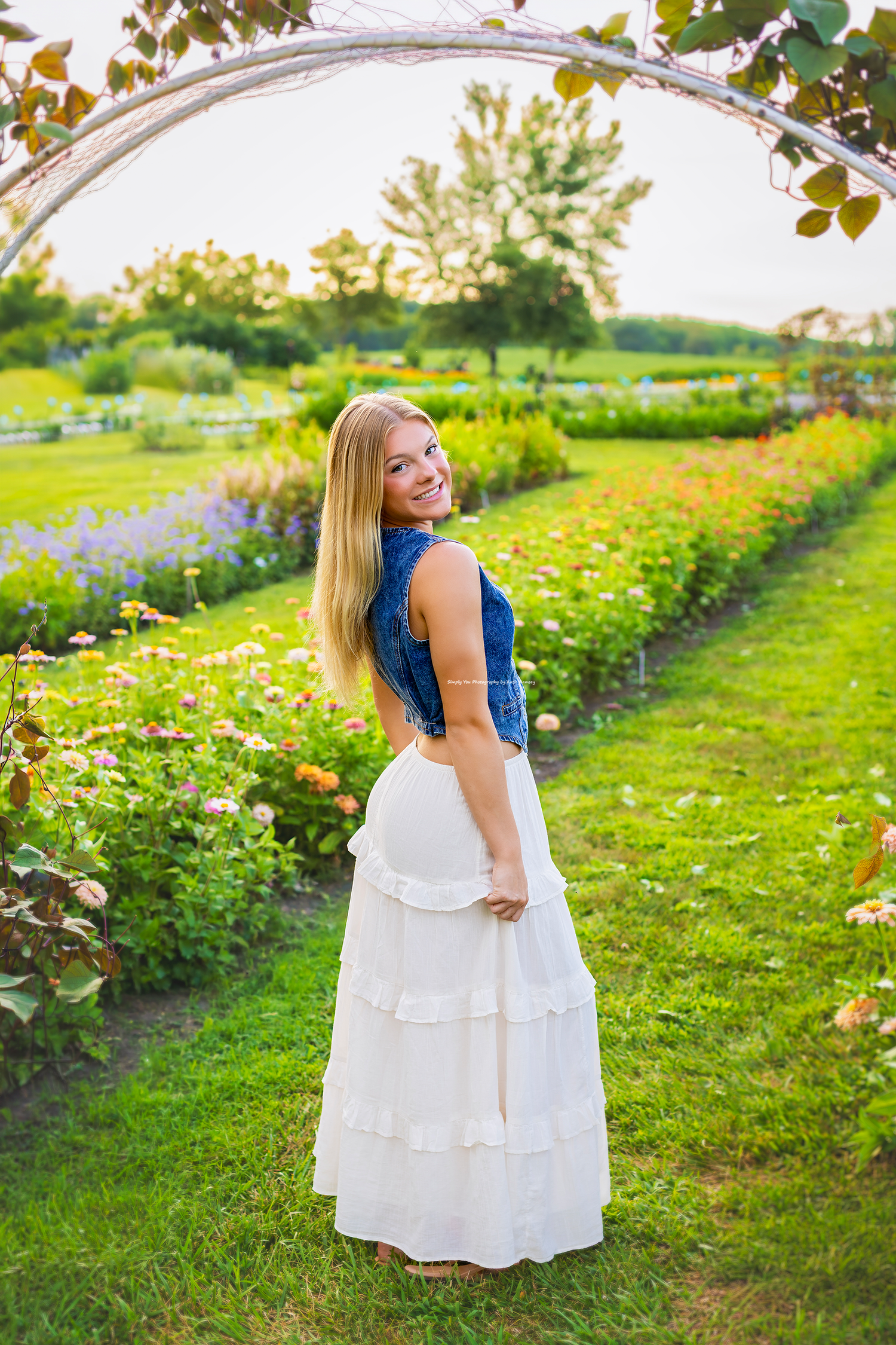 Smiling blonde woman in white tiered skirt and denim top standing under floral arch in colorful garden.