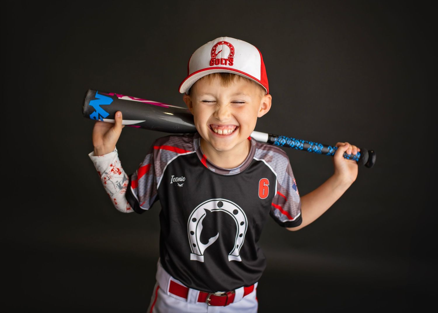 Smiling boy in baseball uniform holds bat over shoulders against dark studio background.