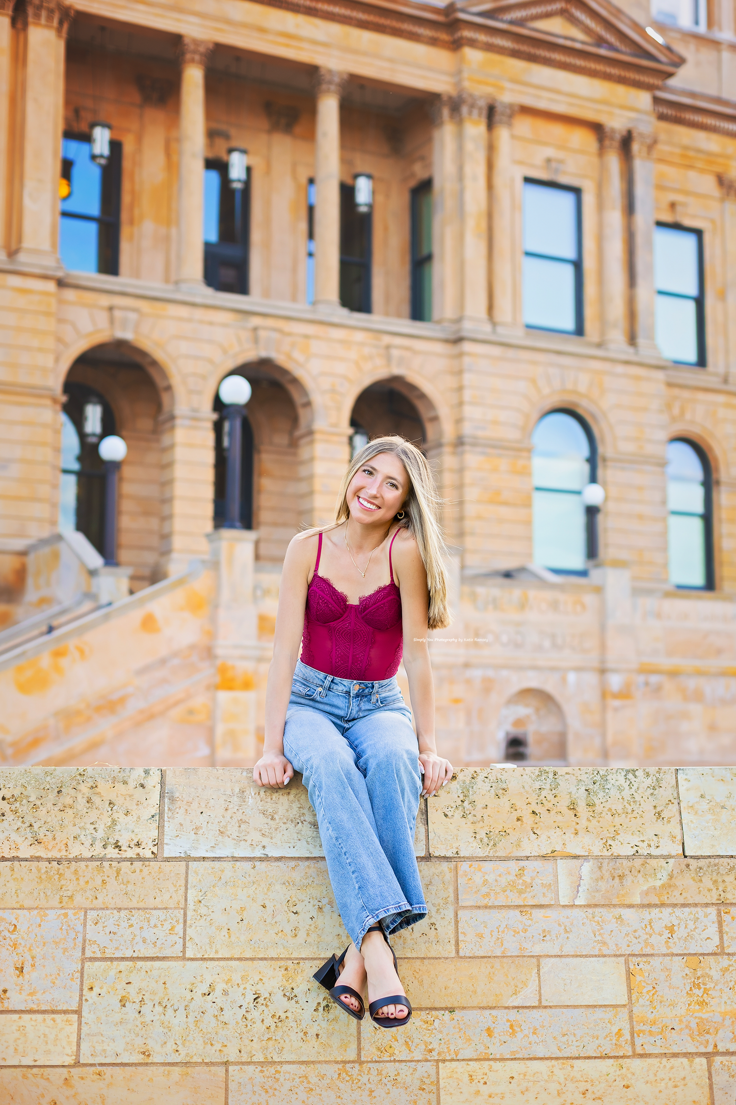Young blonde woman in red top and jeans sitting on stone wall outside classic brick building.