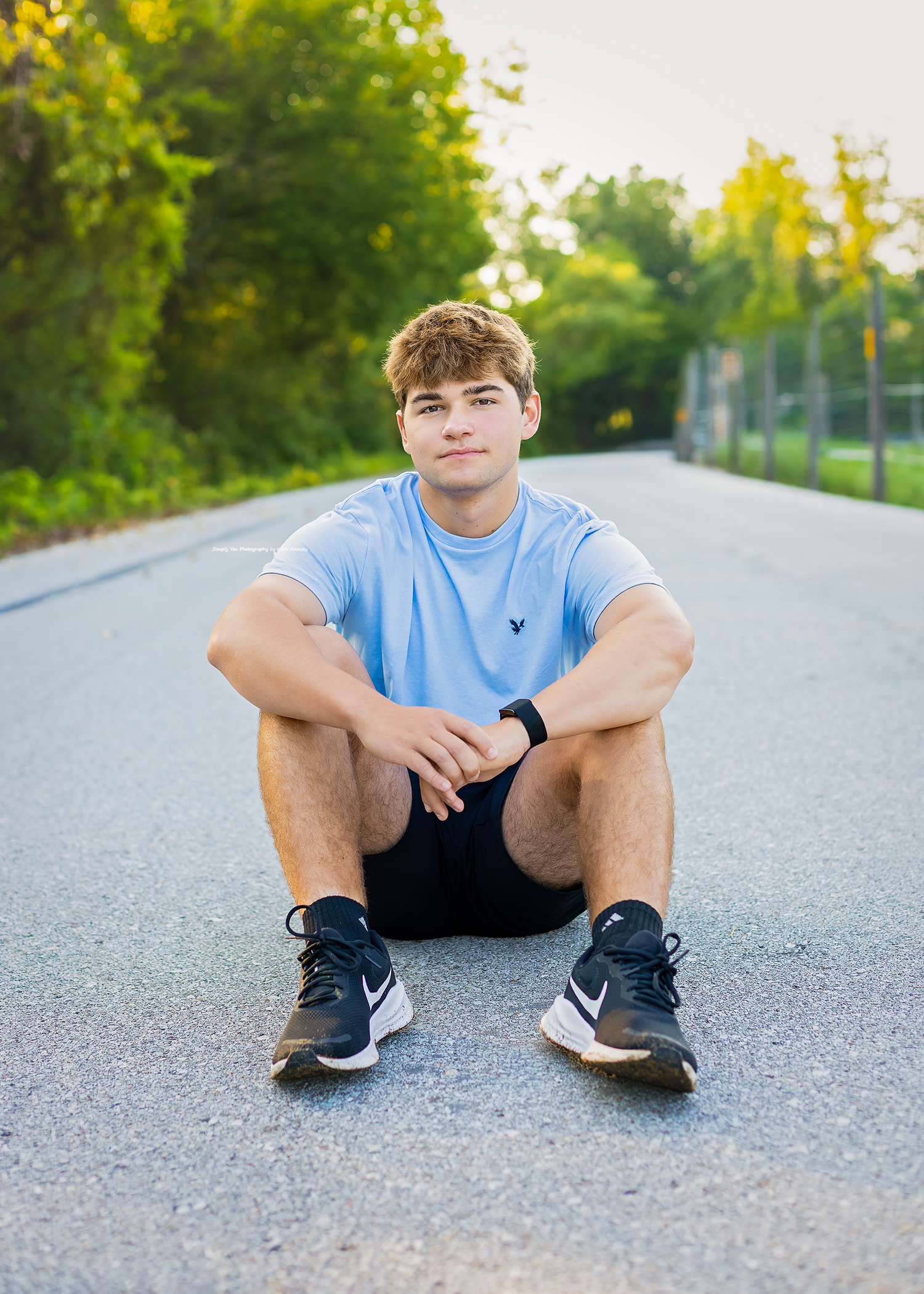 Teen boy in light blue shirt and Nike shoes sits on a road path surrounded by green trees in golden hour light.