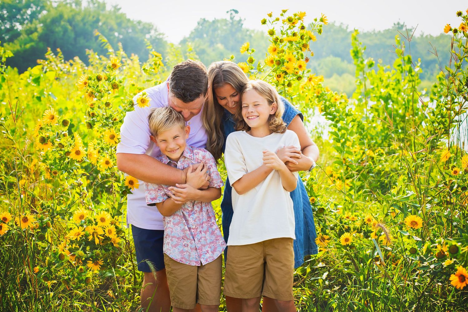 Happy family of four laughing together among vibrant yellow wildflowers in a sunny field.
