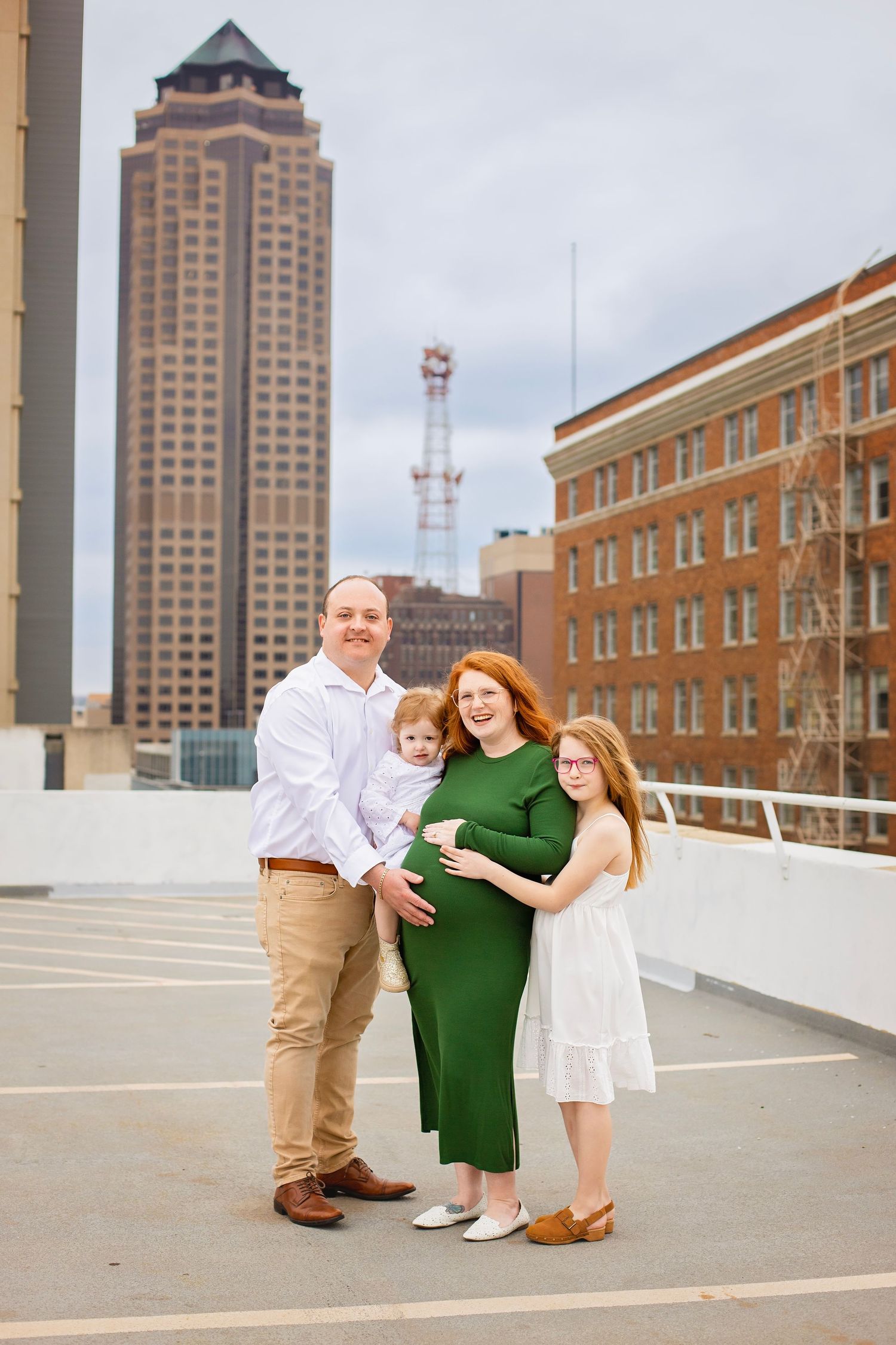 Family of four with pregnant redhead mom in green dress on urban rooftop with city buildings.