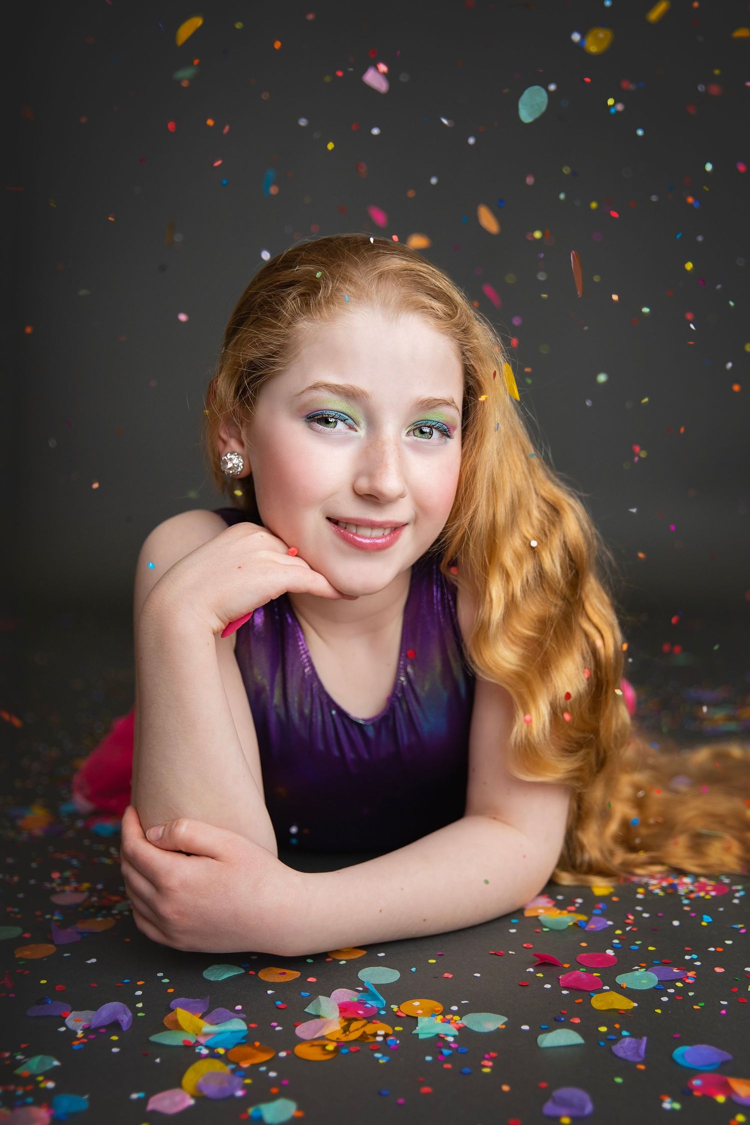A smiling young girl with long red hair in a purple top lies among colorful confetti on a dark background.