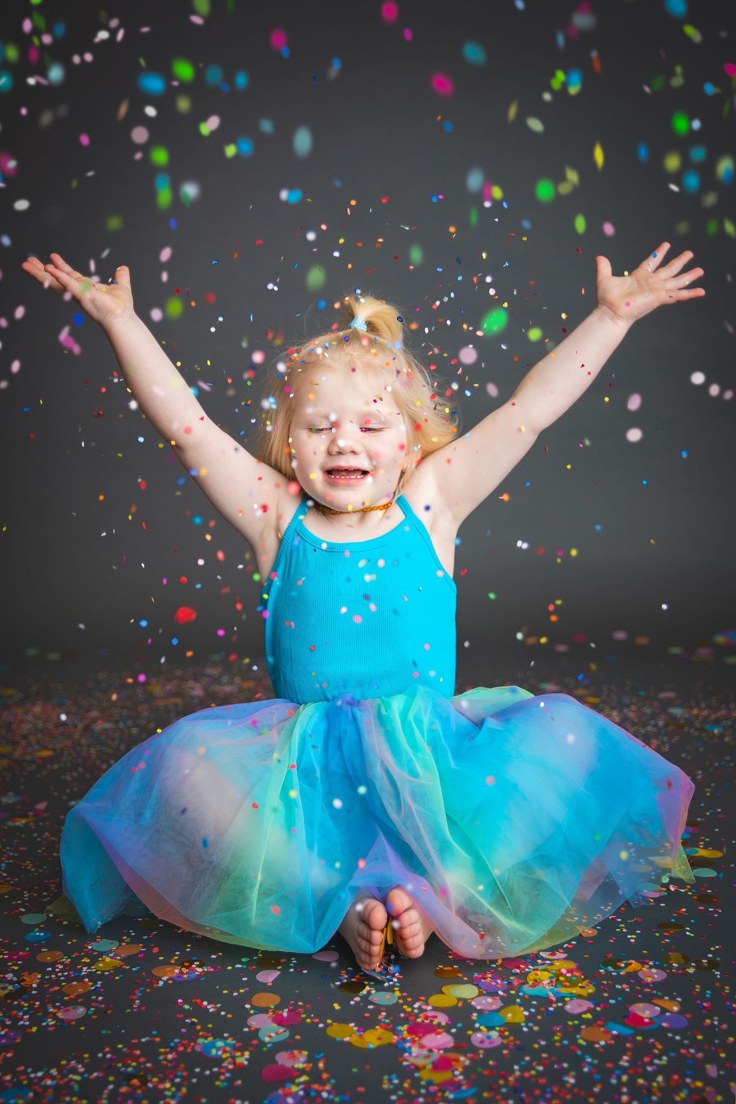Joyful toddler in blue rainbow tutu celebrating with arms raised as colorful confetti falls.