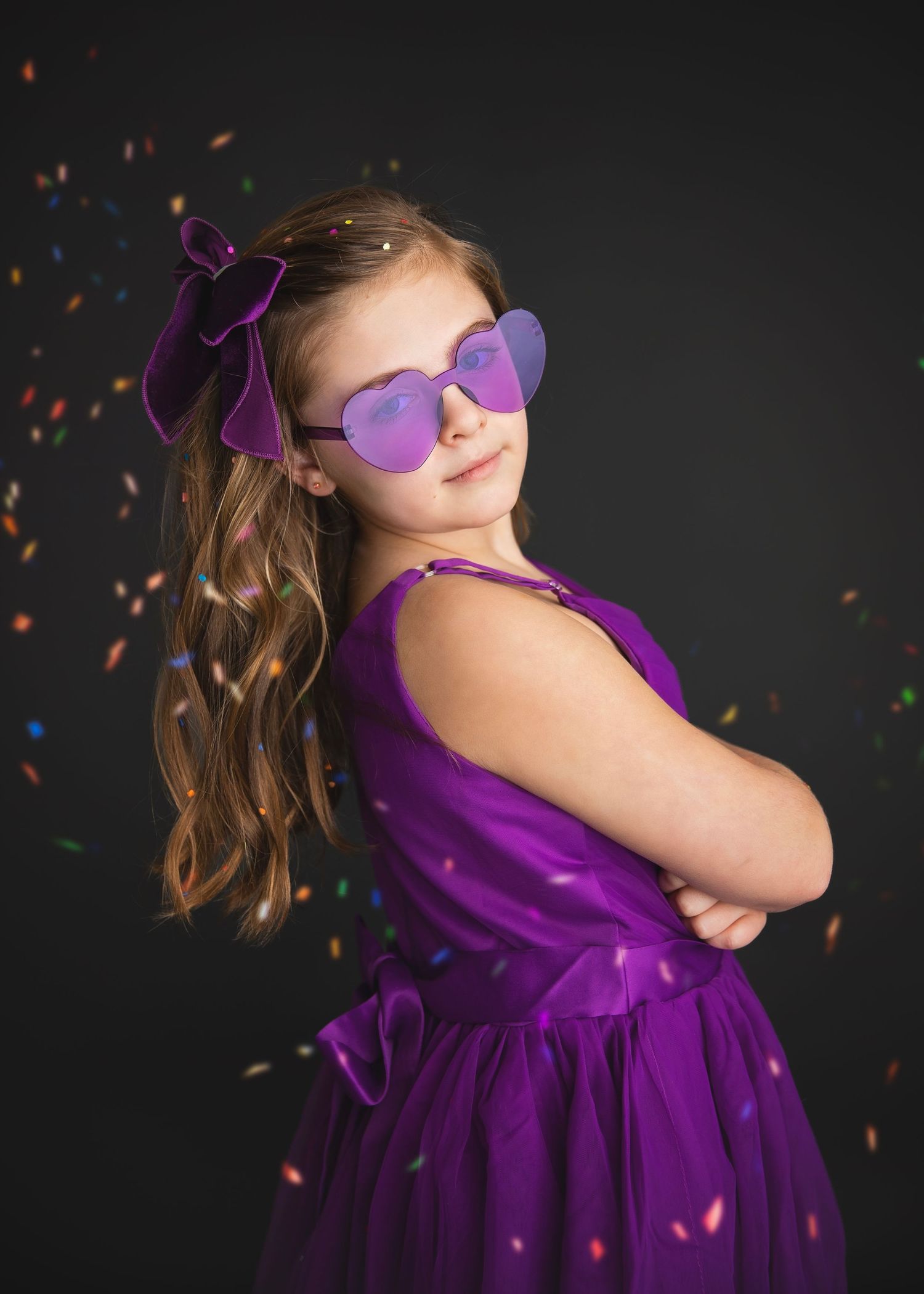 Stylish girl in purple dress and heart-shaped sunglasses posing confidently with arms crossed amid confetti.