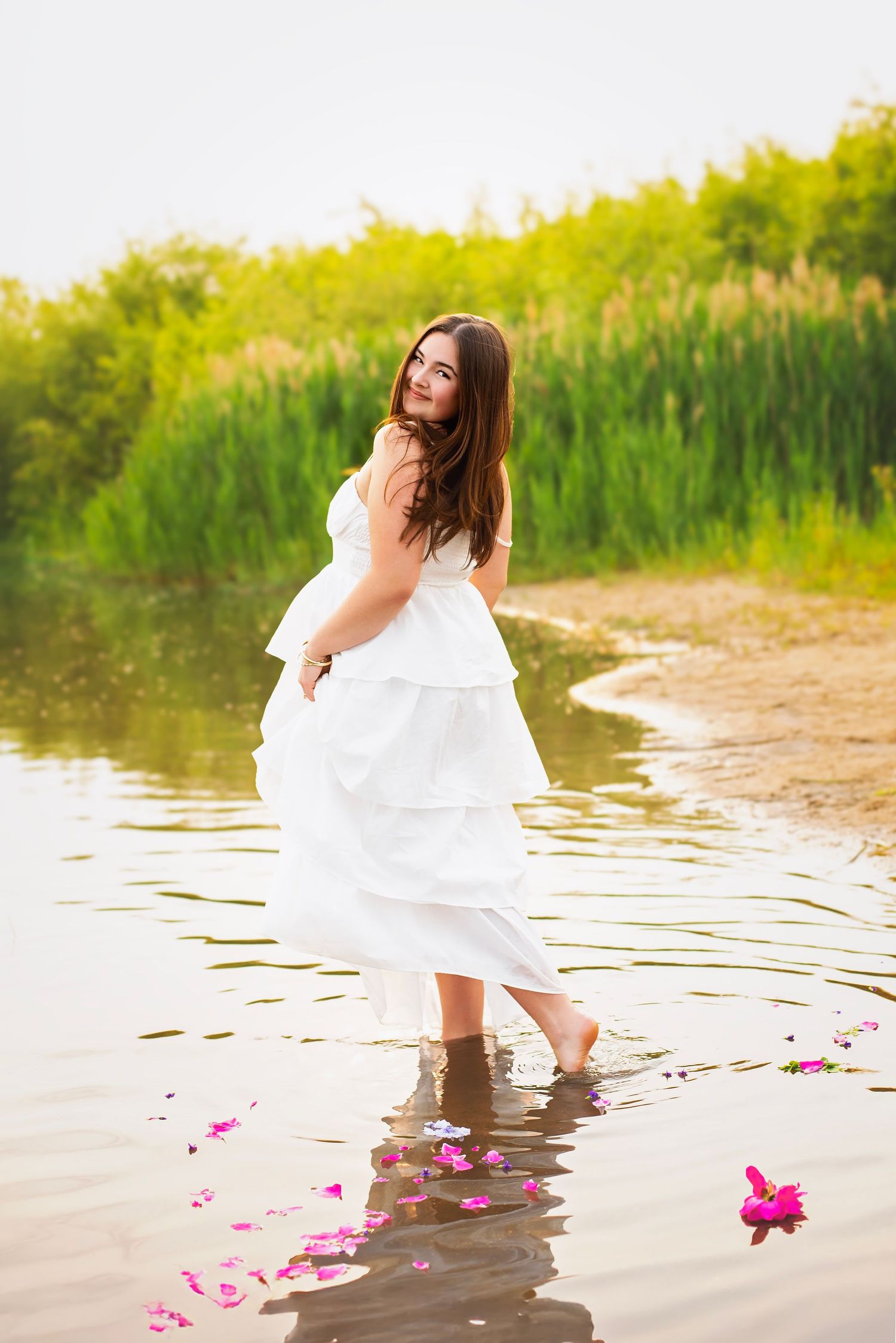 Young woman in white dress wading in shallow water with pink flower petals floating nearby.