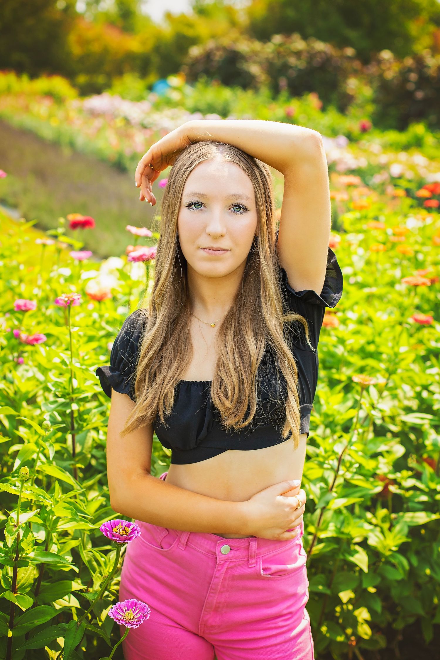 Blonde teen girl in black crop top and pink jeans posing in colorful flower garden.