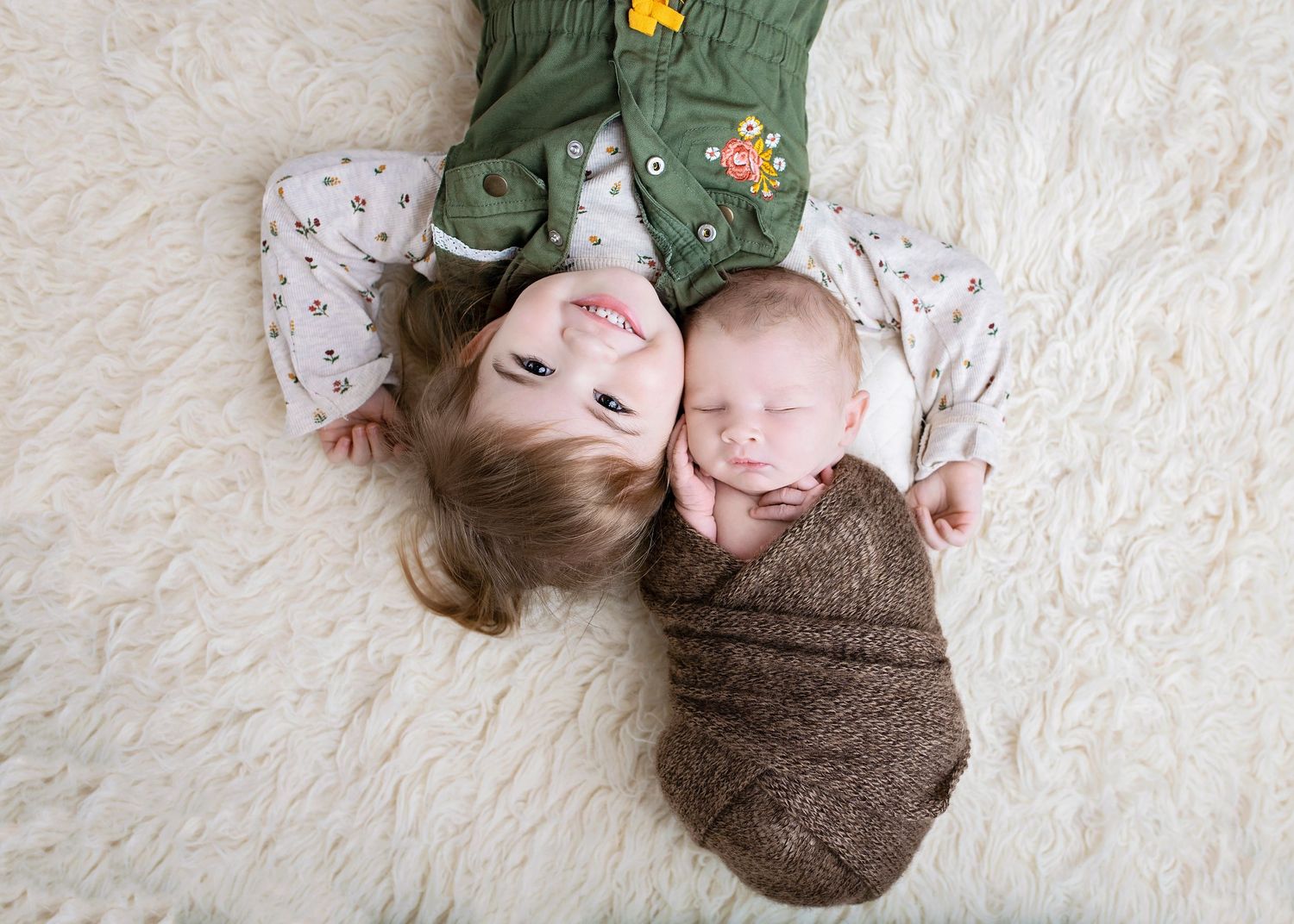 Young child and swaddled newborn baby lying together on a soft cream fur rug, viewed from above.
