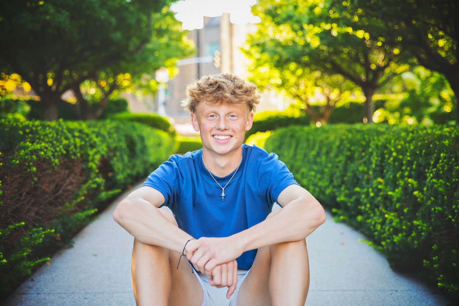 Smiling teen boy with curly blonde hair sits on garden path surrounded by lush green hedges.