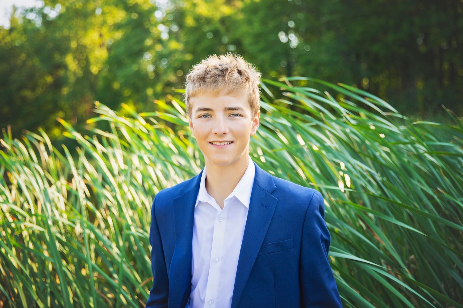 Smiling teenage boy in a blue suit jacket posing in front of tall green reeds.
