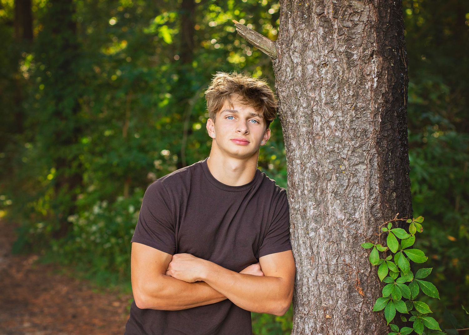 Young man with blond hair leans against a tree in a forest, arms crossed, wearing a dark brown t-shirt.