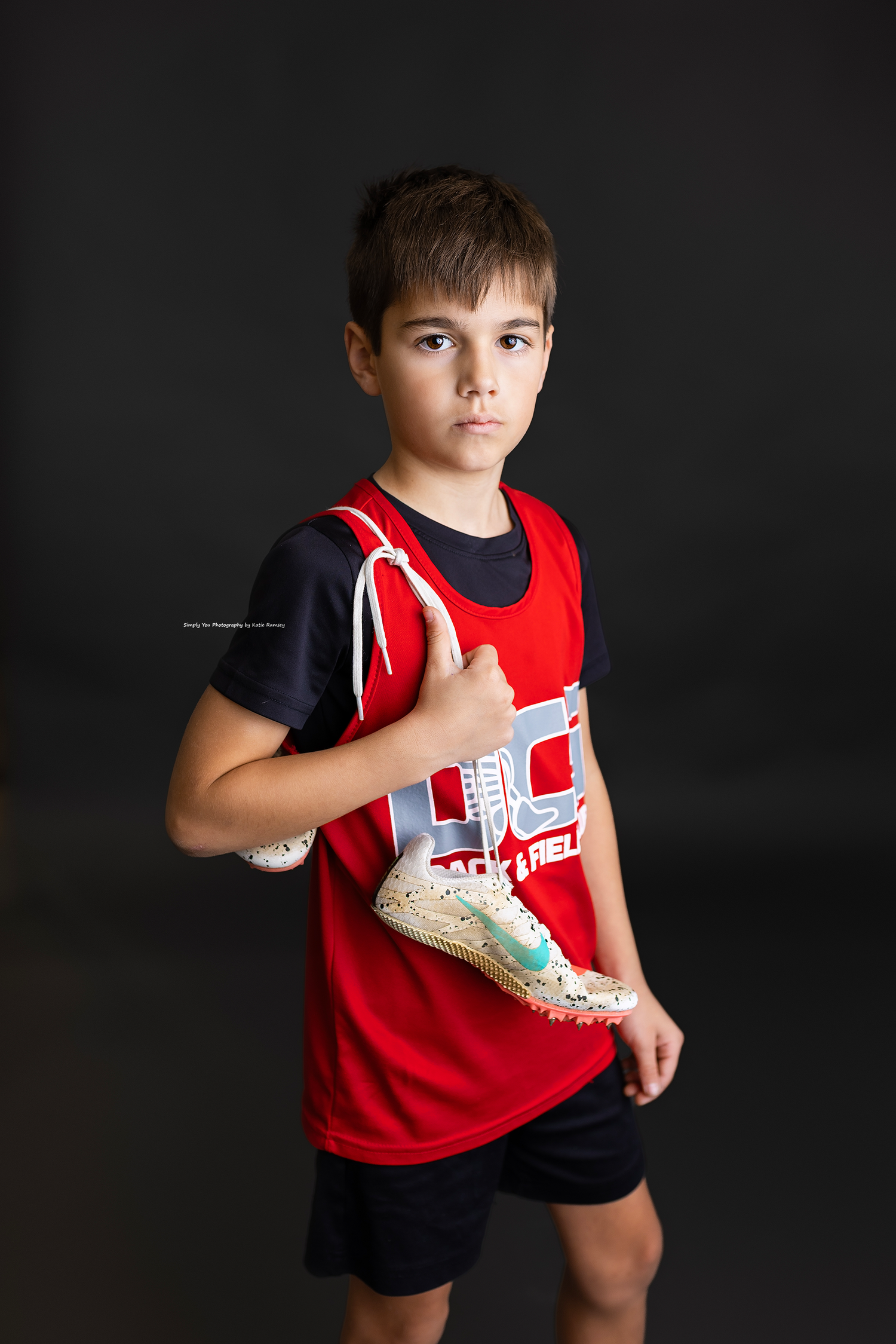 Young boy in red track and field jersey holds athletic spikes shoes against dark studio background.