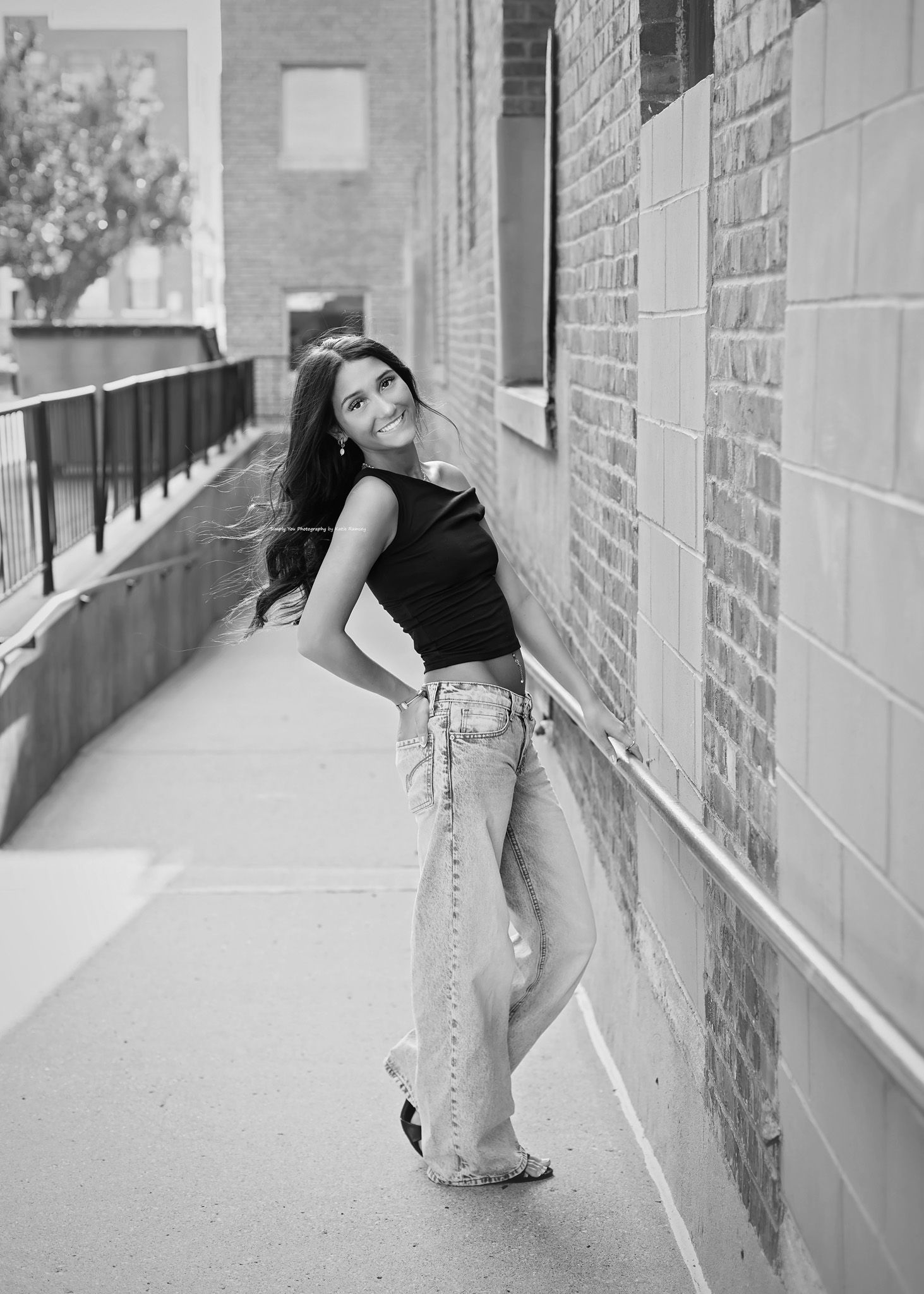 Black and white photo of a young woman in wide-leg jeans and black crop top posing against a brick wall on a sidewalk.