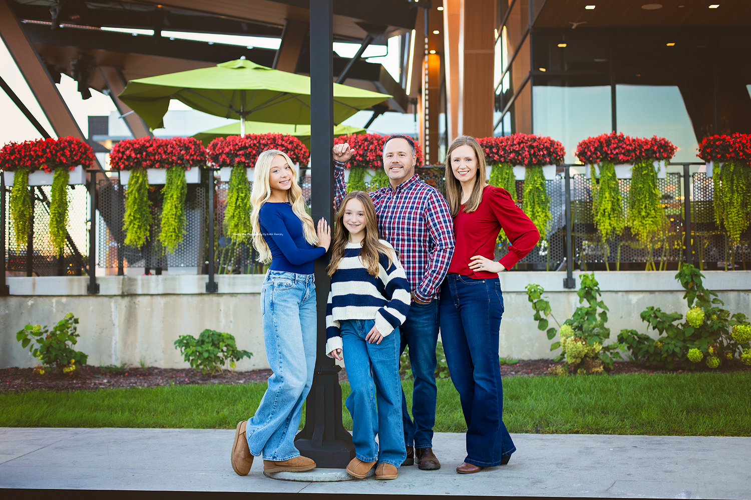 A smiling family of four poses outdoors near festive red floral displays and modern architecture.