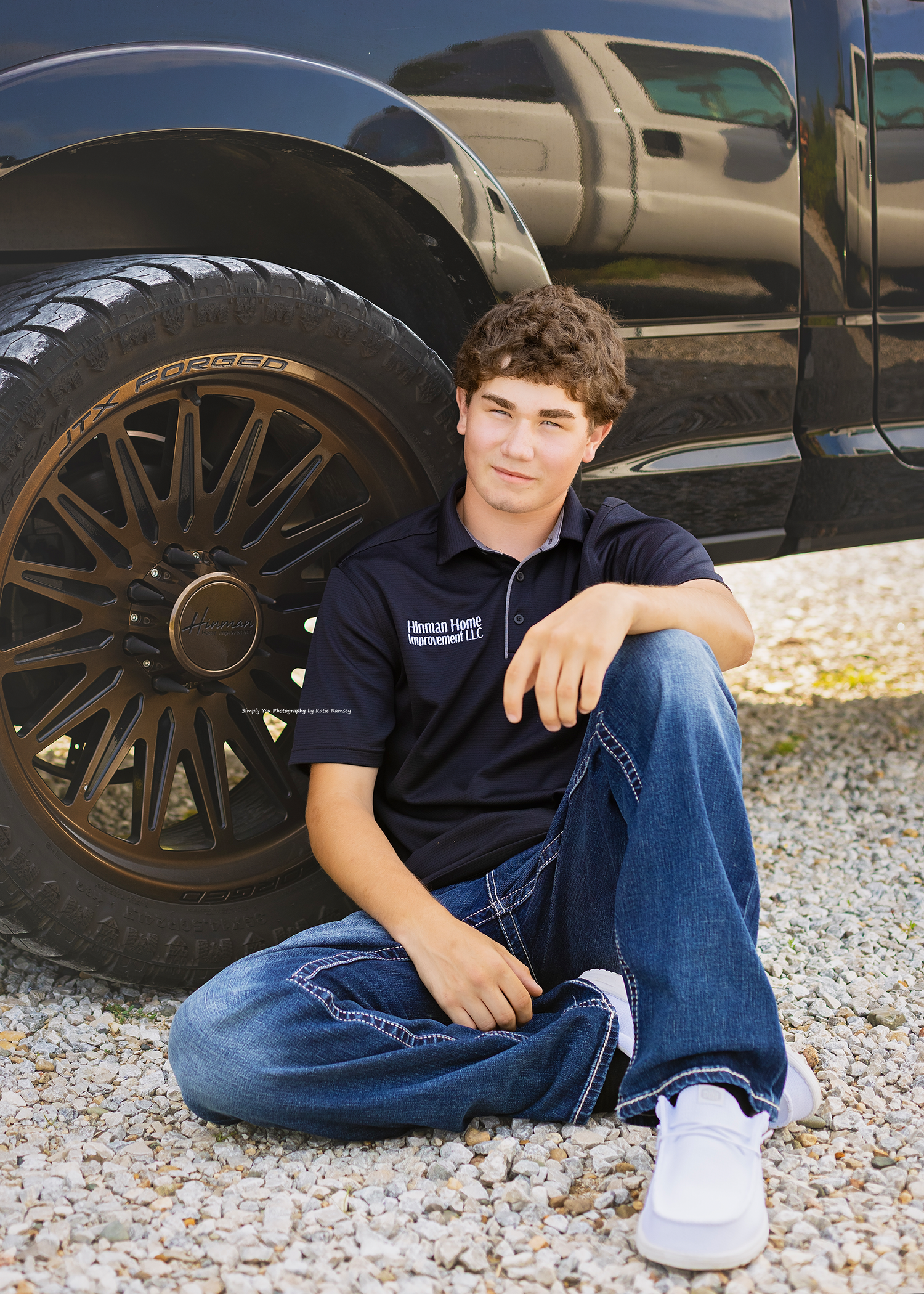 Teen boy in navy polo and jeans sits on gravel leaning against a black truck with dark chrome wheels.