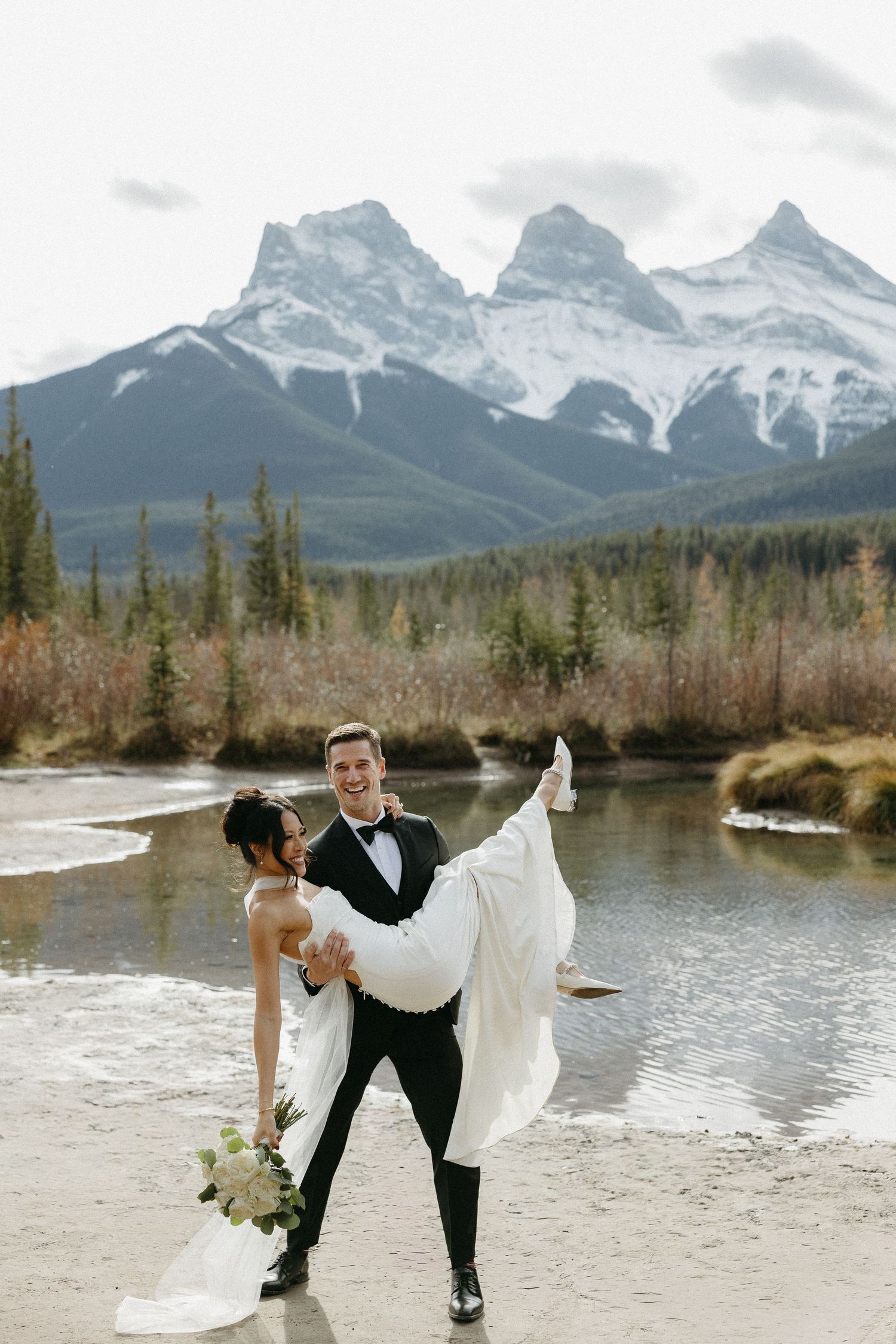 Groom carries bride in wedding attire by a mountain river with snowy peaks in Canmore, Alberta.