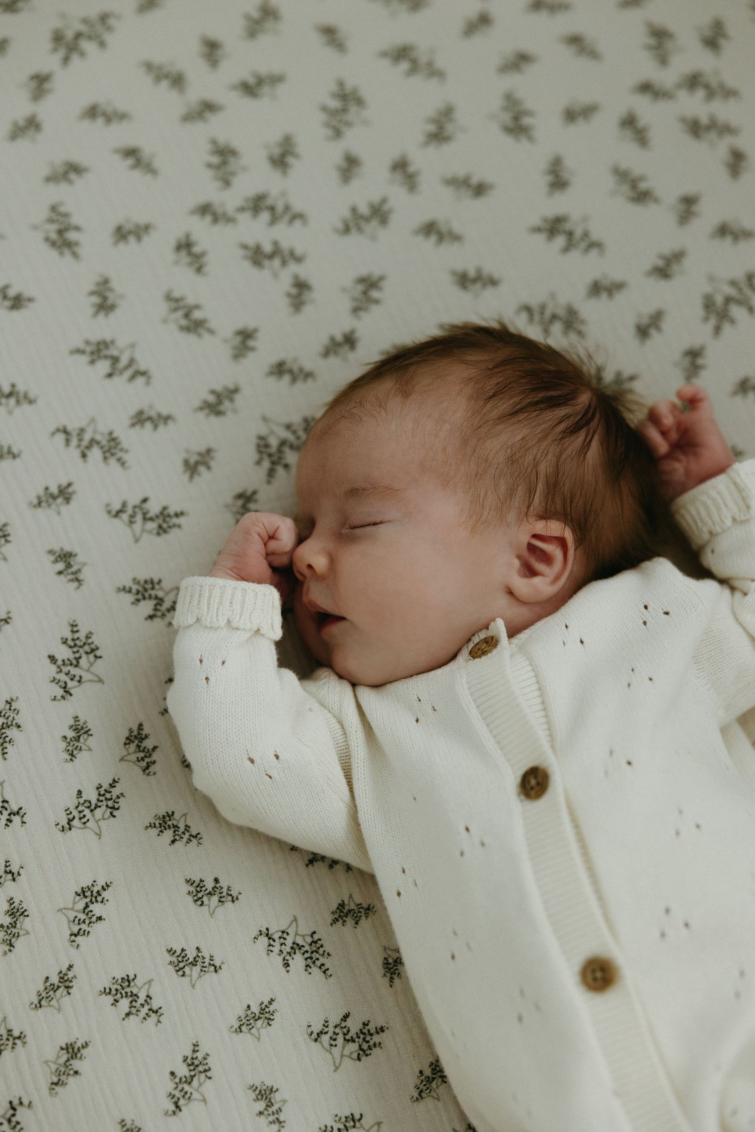 Sleeping infant wearing white knit sweater on floral patterned fabric.