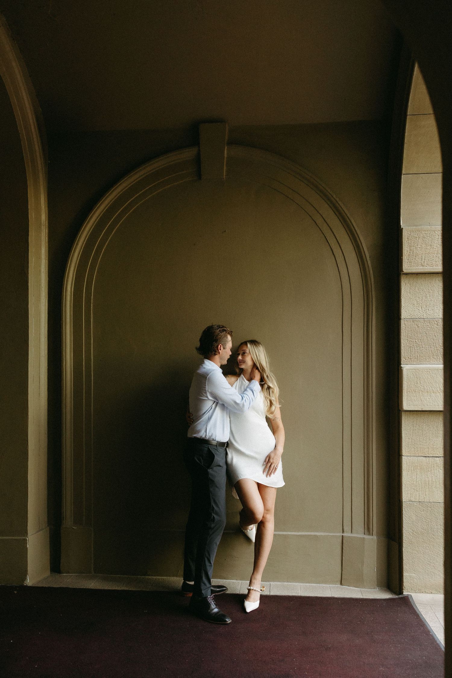 Couple embraces romantically in an arched hallway with dramatic lighting and architectural details.