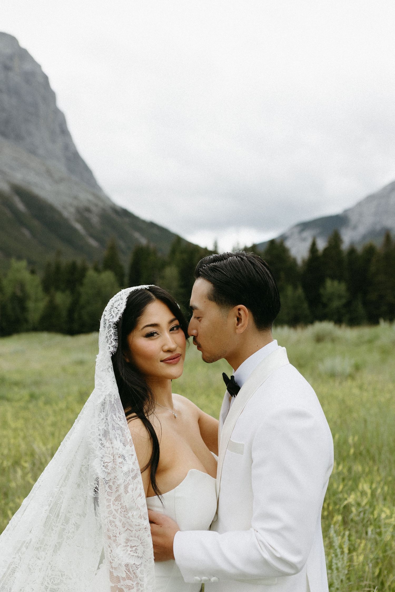 A couple in wedding attire poses in a mountain meadow, the groom in white kissing the bride's temple as she smiles.